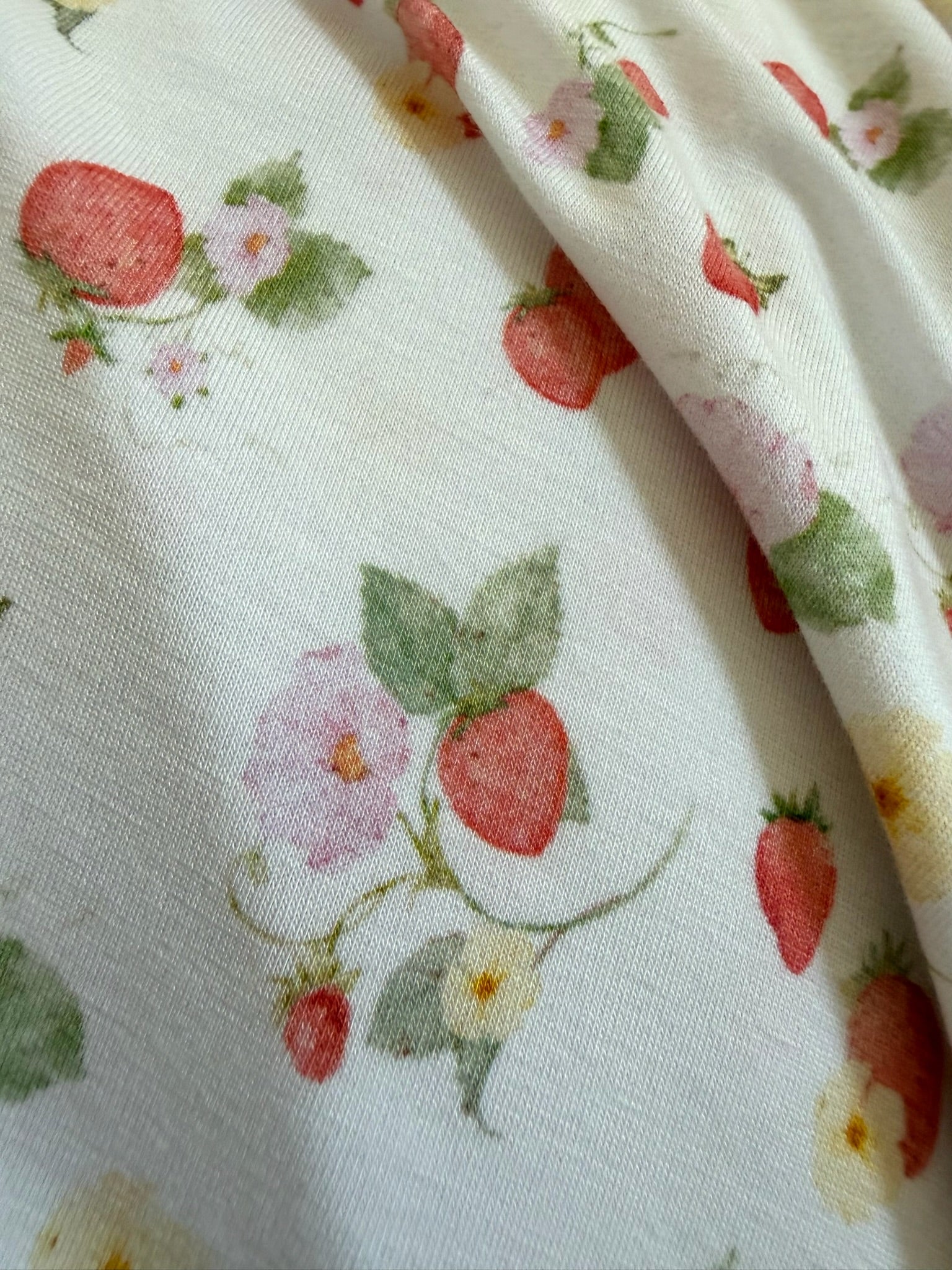 Close-up of "freshly picked" fabric: white base with a garden strawberry print—red strawberries, green leaves, and small pink and yellow flowers. Gentle folds and shadows form in the slightly wrinkled fabric.