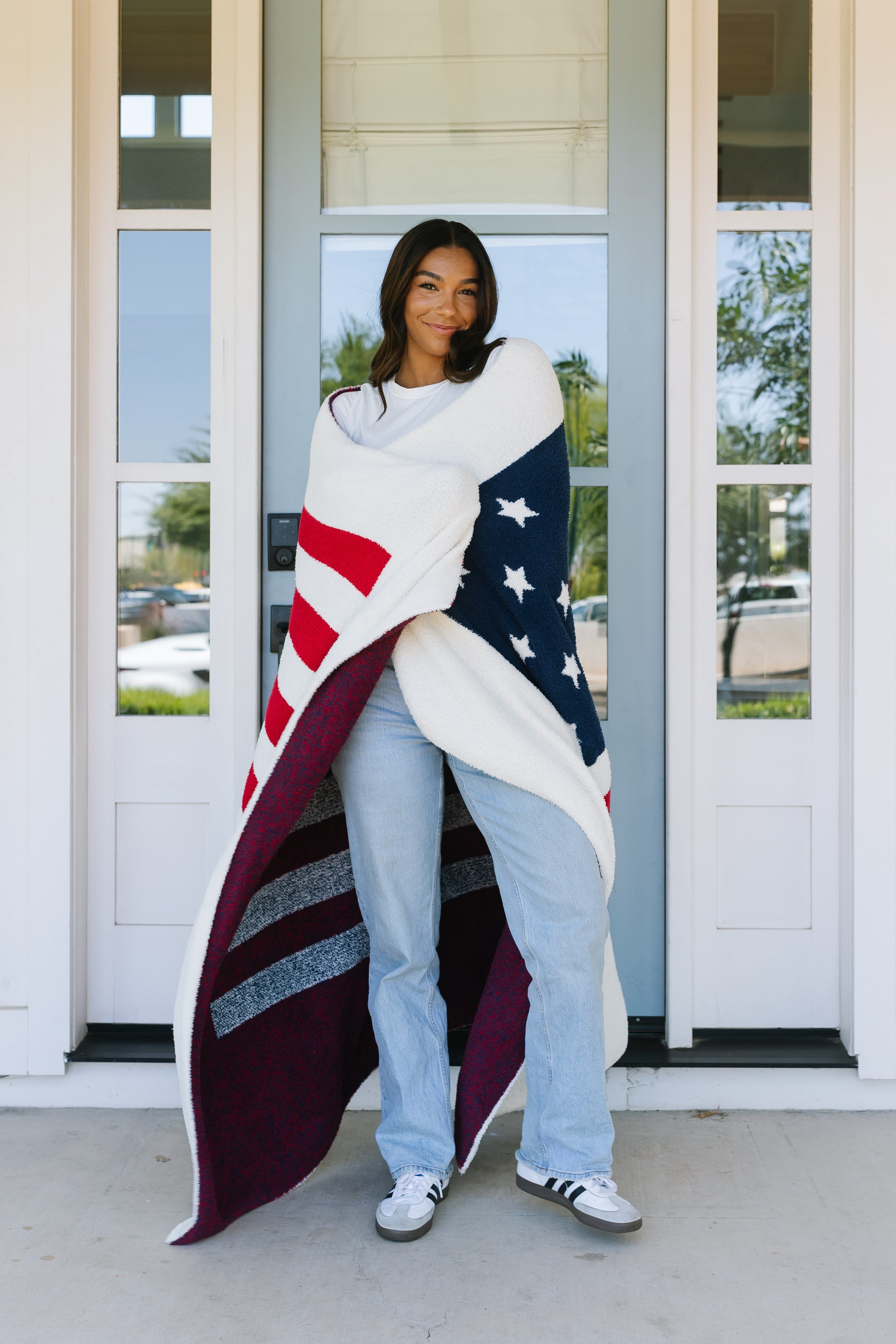 A woman smiles on a porch in front of a light blue door, wrapped in the america / plush throw designed like an American flag. She wears jeans, a white shirt, and sneakers; house number 229 is visible nearby.
