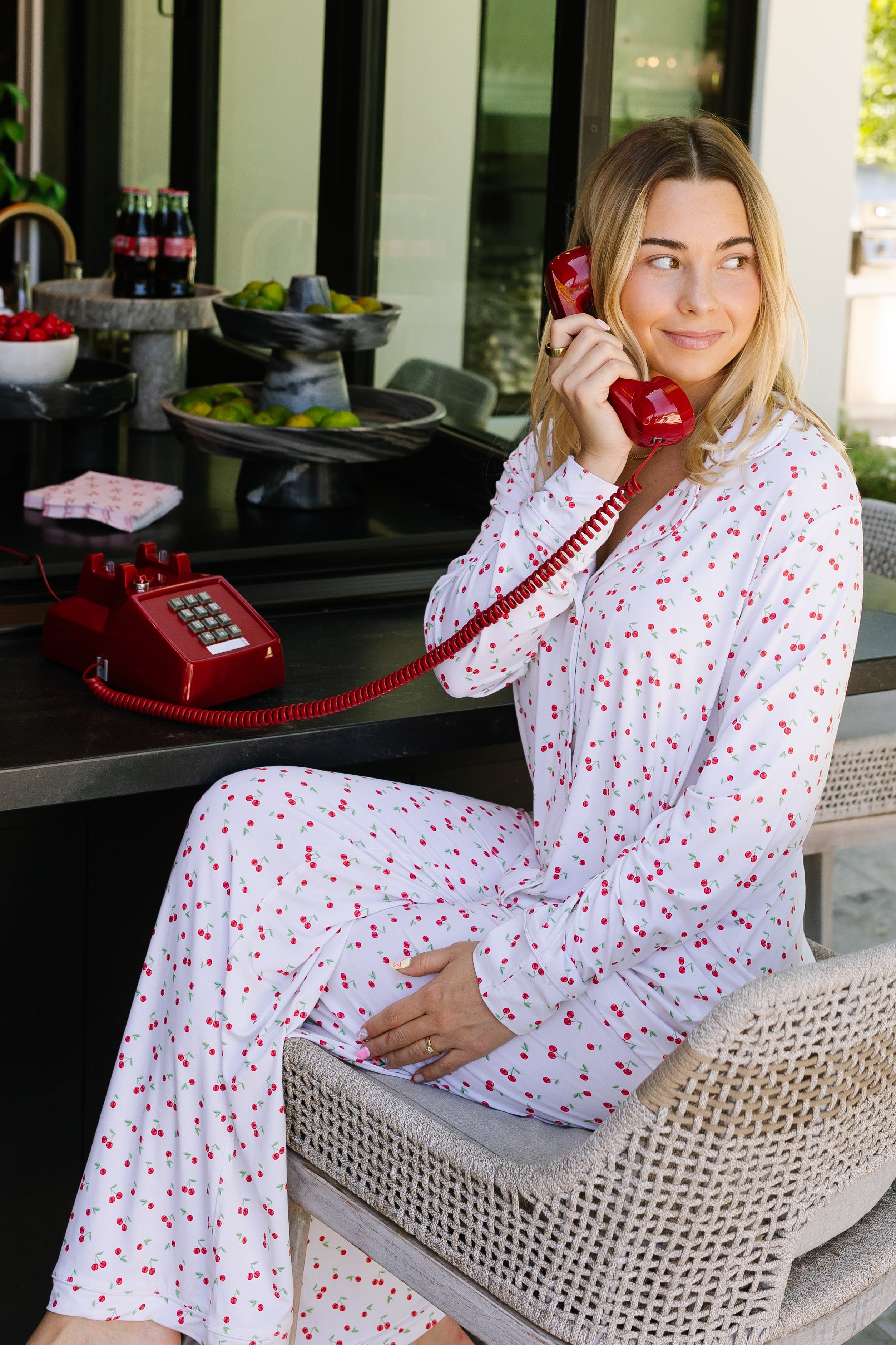 Wearing the cherry on top pajama set with red hearts, a woman smiles at an outdoor table, holding a red vintage phone. Drinks and snacks sit nearby, with greenery and a white house in the background.