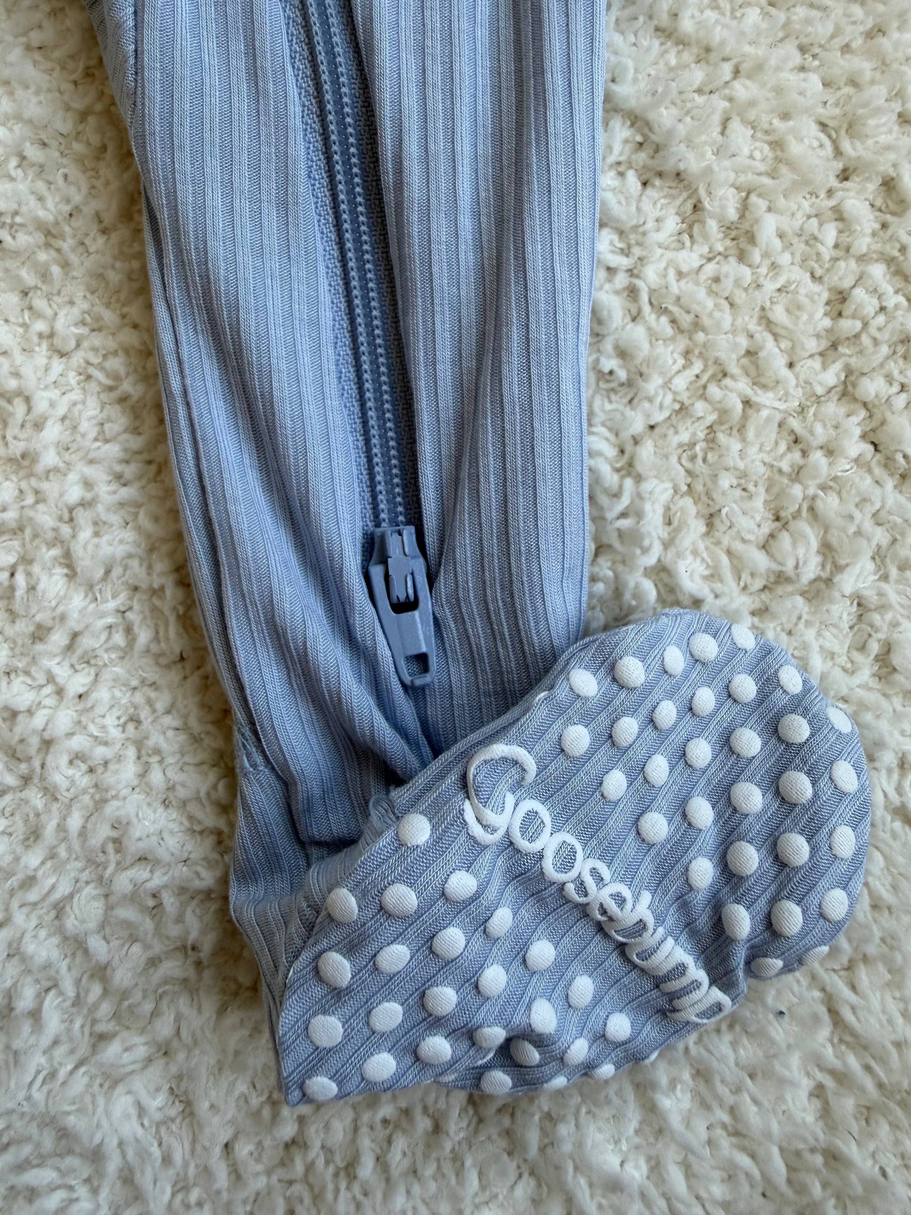 A close-up of the cove / one piece zipper shows a blue, ribbed bamboo baby sleeper with a zipper and white grip dots on the sole. The word Goosebumps is visible on the bottom of the foot. These cozy footed pajamas are lying on a textured, cream-colored carpet.