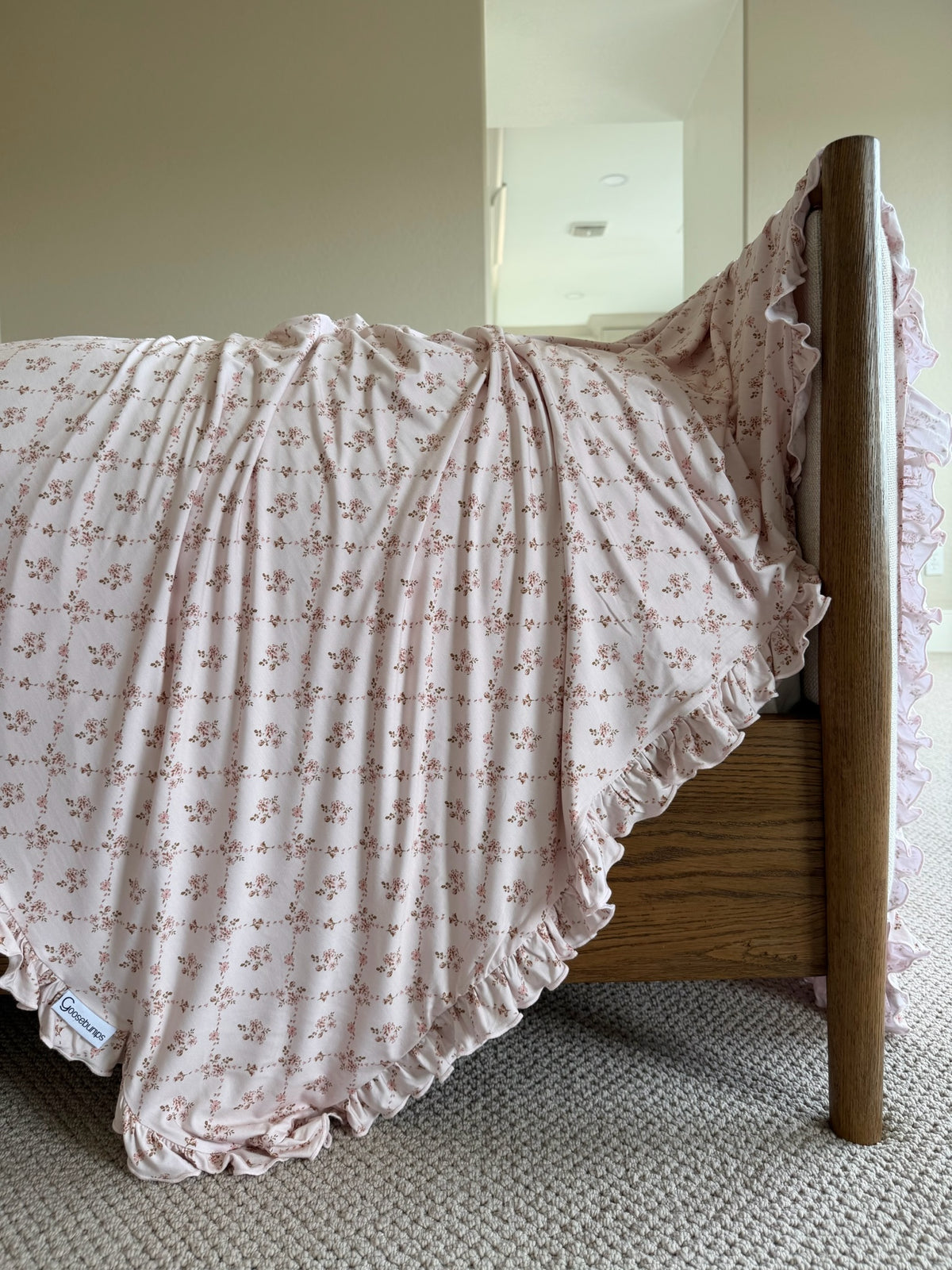 A keepsake / ruffle bed showcases a luxurious floral blanket with pink ruffled edges, elegantly draped over the side of its wooden frame, resting on a light carpeted floor in a softly lit, neutral-toned room.