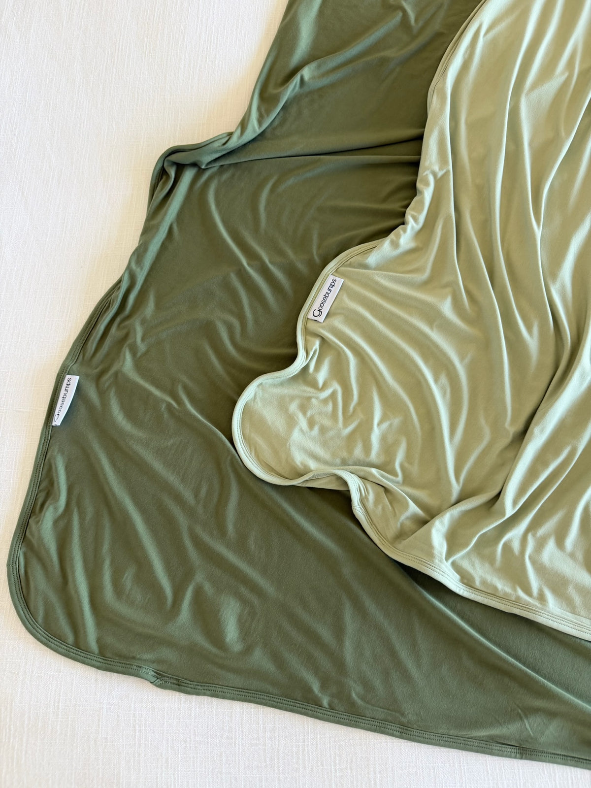 Two winter pine bamboo blankets in olive and sage green draped over a white surface, each featuring a brand label on the edge.