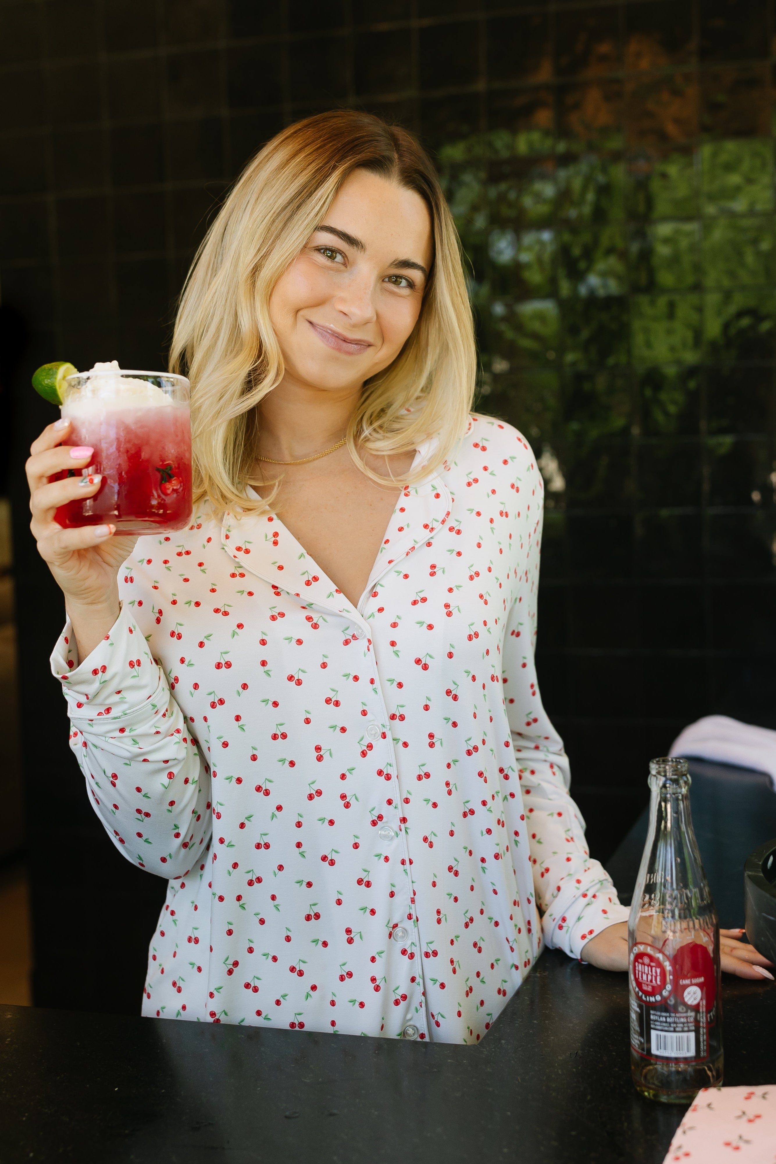 A woman with blonde hair smiles in her kitchen wearing the cherry on top pajama set: a relaxed fit white shirt with red floral patterns. She holds a red drink with ice and lime, with cherries and a glass bottle on the counter before her.