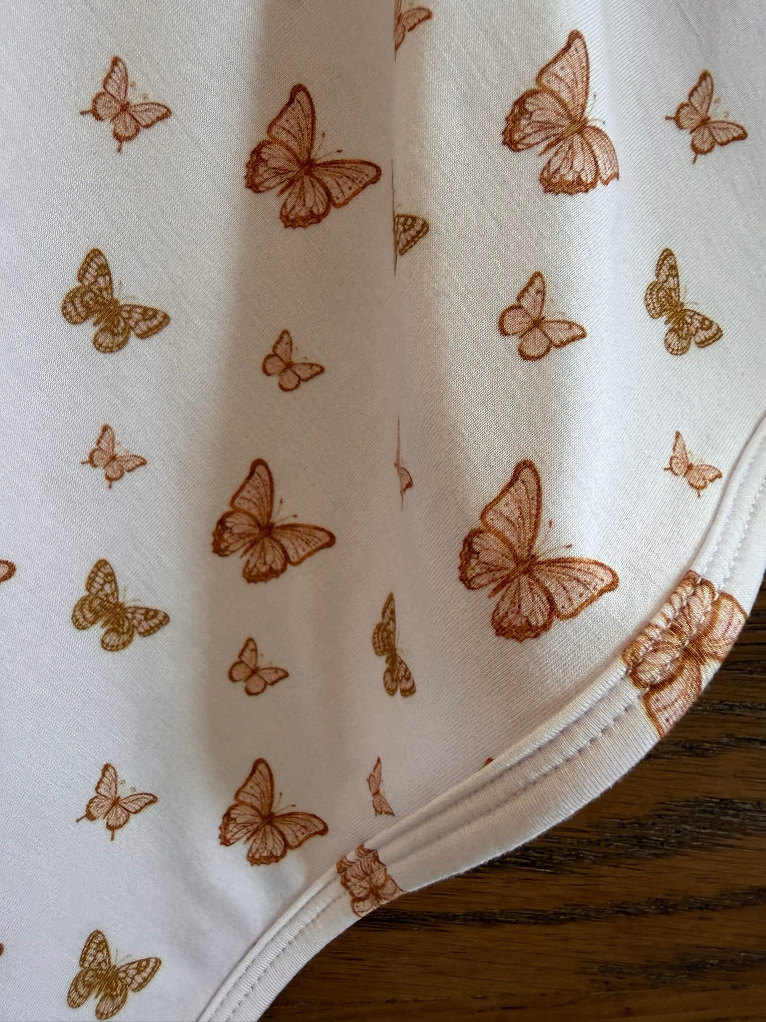 A close-up of flutter, an adult swaddle made with white bamboo fabric and a brown-gold butterfly print of varying sizes. The swaddle is displayed on a wooden surface, showing the edge seams.