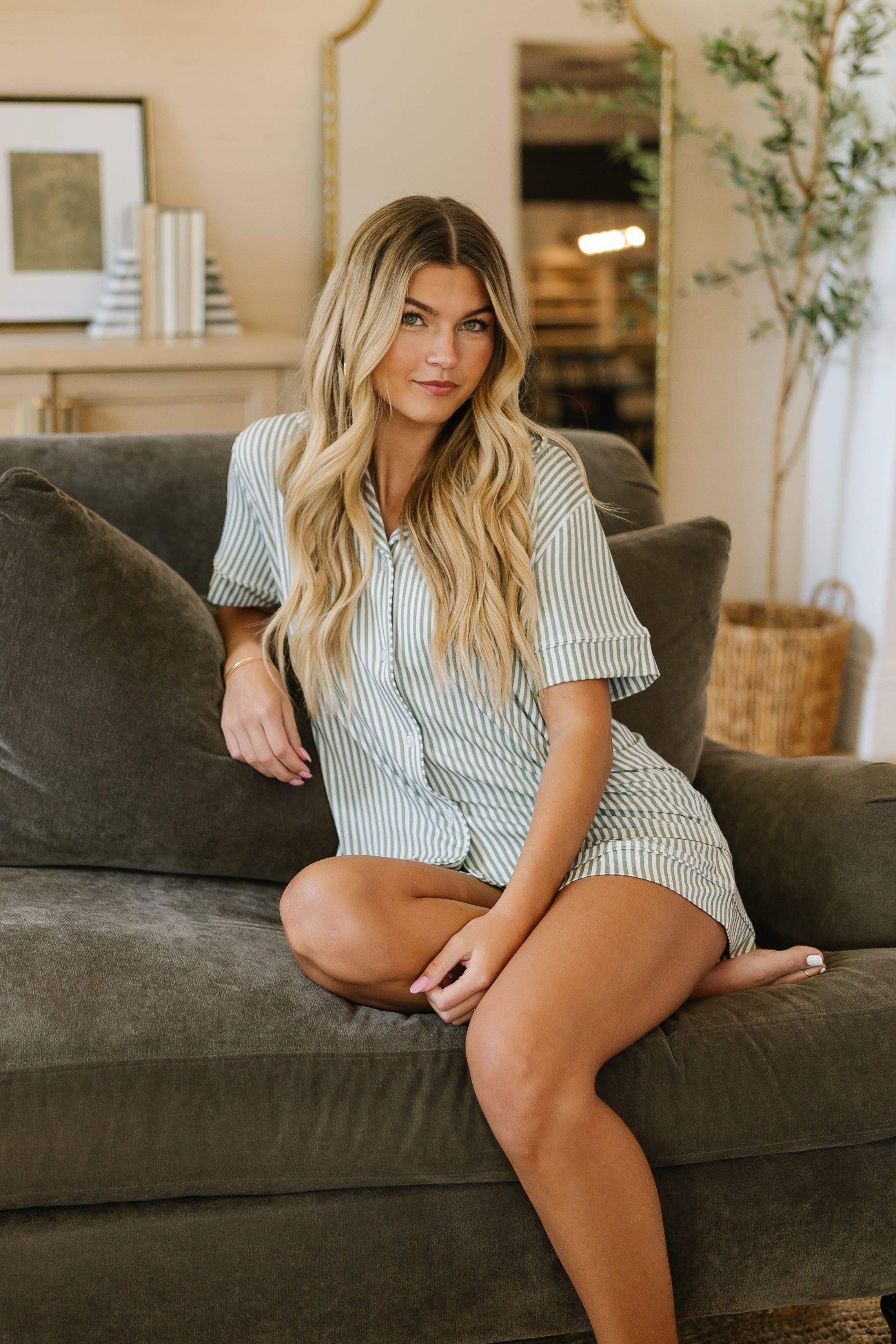 A woman with long blonde hair sits barefoot on a dark gray sofa in a modern living room, wearing the tree house pajama set with shorts. Light walls, chic shelves, and a large mirror create an inviting spring vibe.