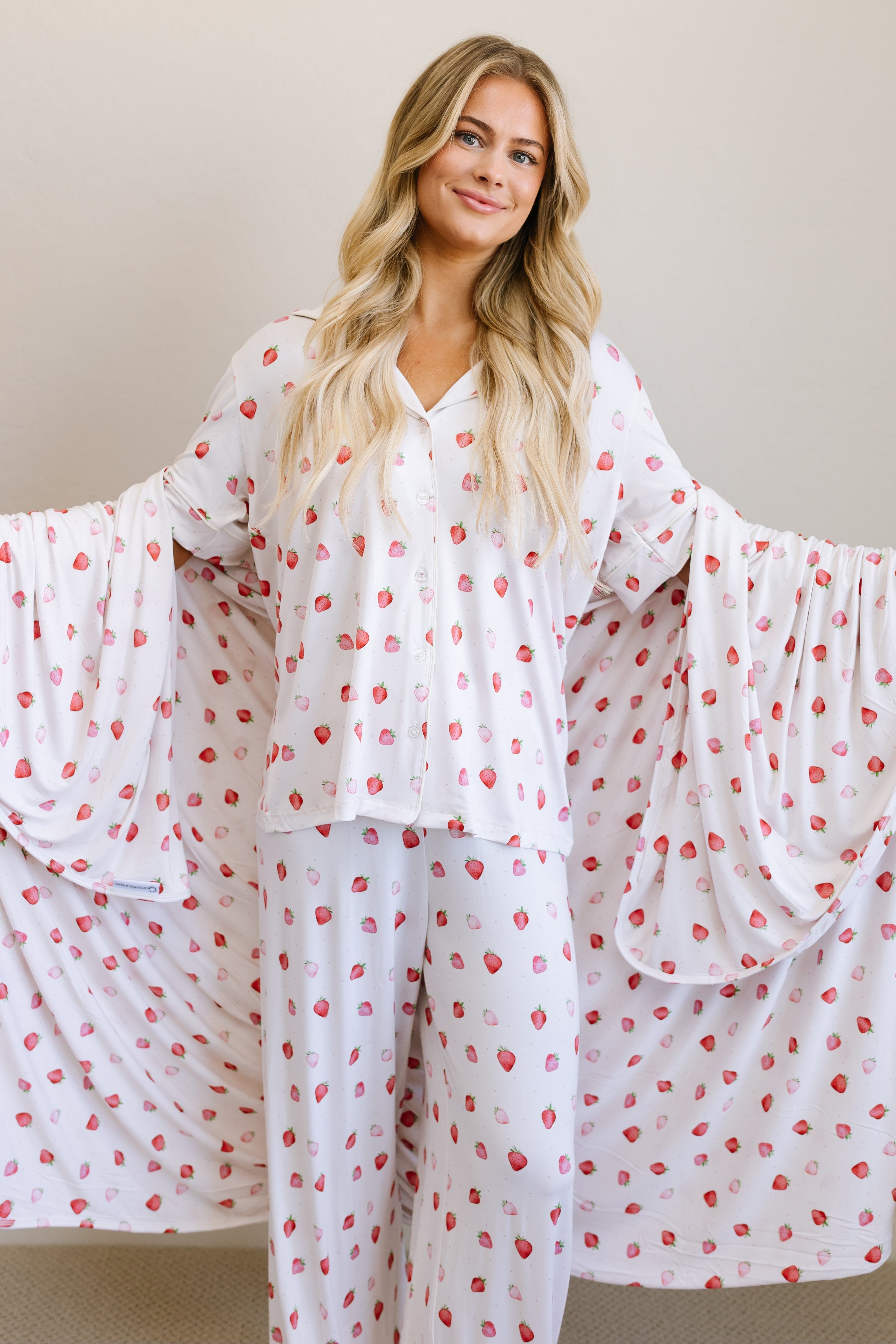 A woman with long blonde hair wears the "strawberry shortcake" white pajamas with a red and pink strawberry pattern, holding a matching strawberry print blanket draped behind her like wings against a plain light background.