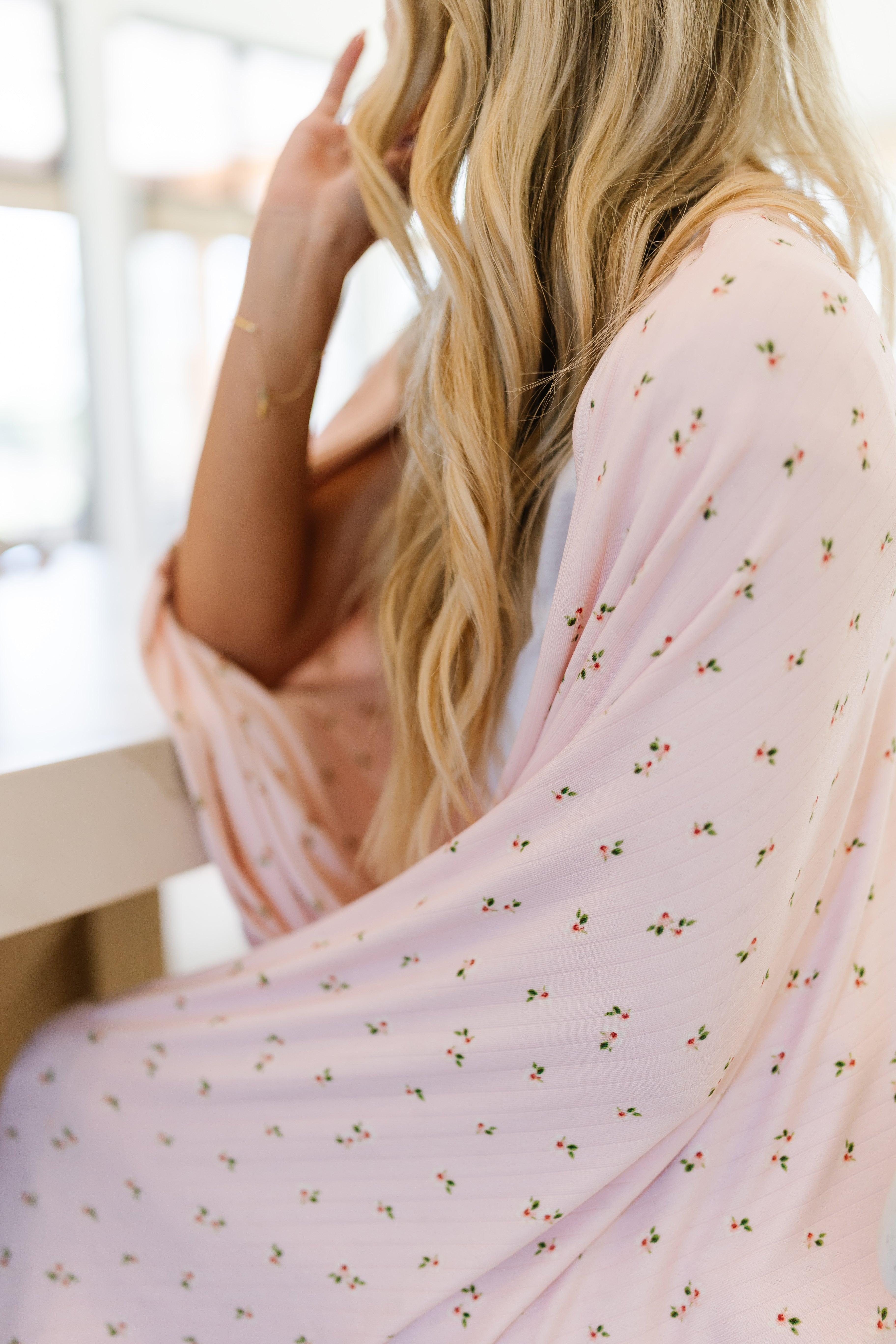 A woman with long, wavy blonde hair sits at a table, draped in the whimsical pink pointelle blanket. Her face is partially out of view, and she rests her hand near her cheek.