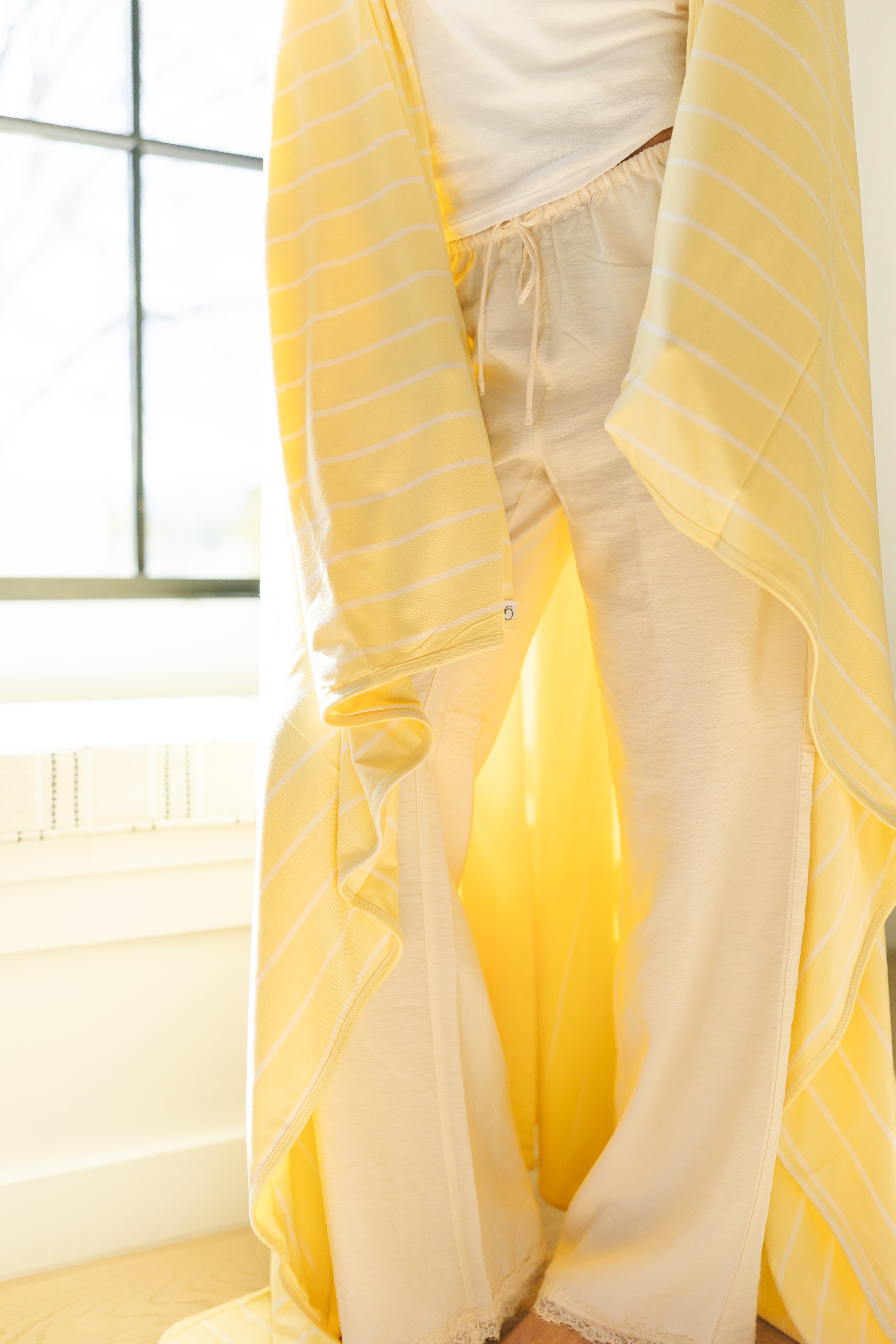 Wrapped in the luxurious lazy days blanket, a person stands by a sunlit window wearing cream pajama pants, a white top, and a long yellow striped robe that drapes to the floor.