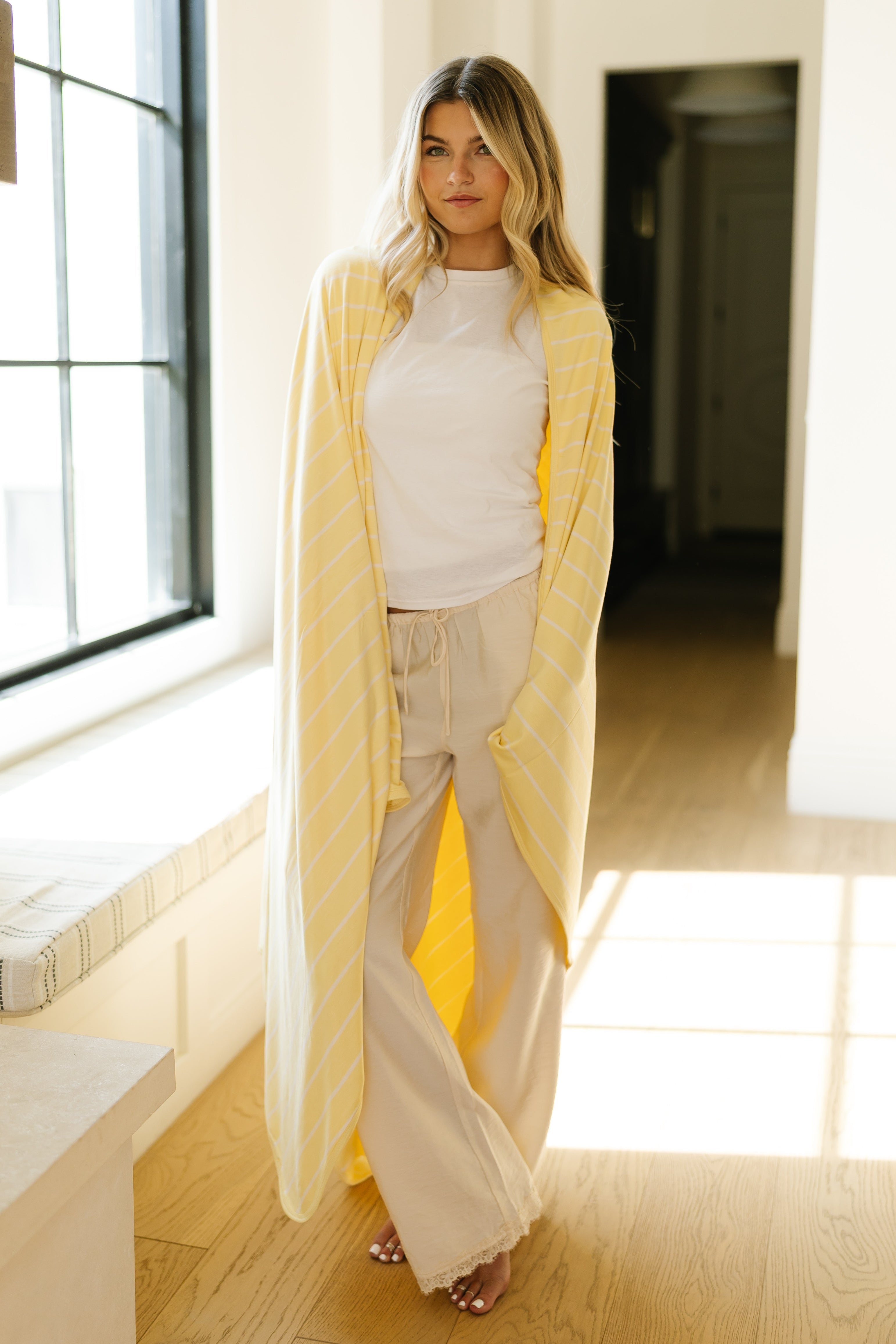 A woman stands indoors on a wooden floor, sunlight pouring in through a large window, wearing beige pants, a white t-shirt, and the "lazy days" blanket with yellow stripes draped over her shoulders.