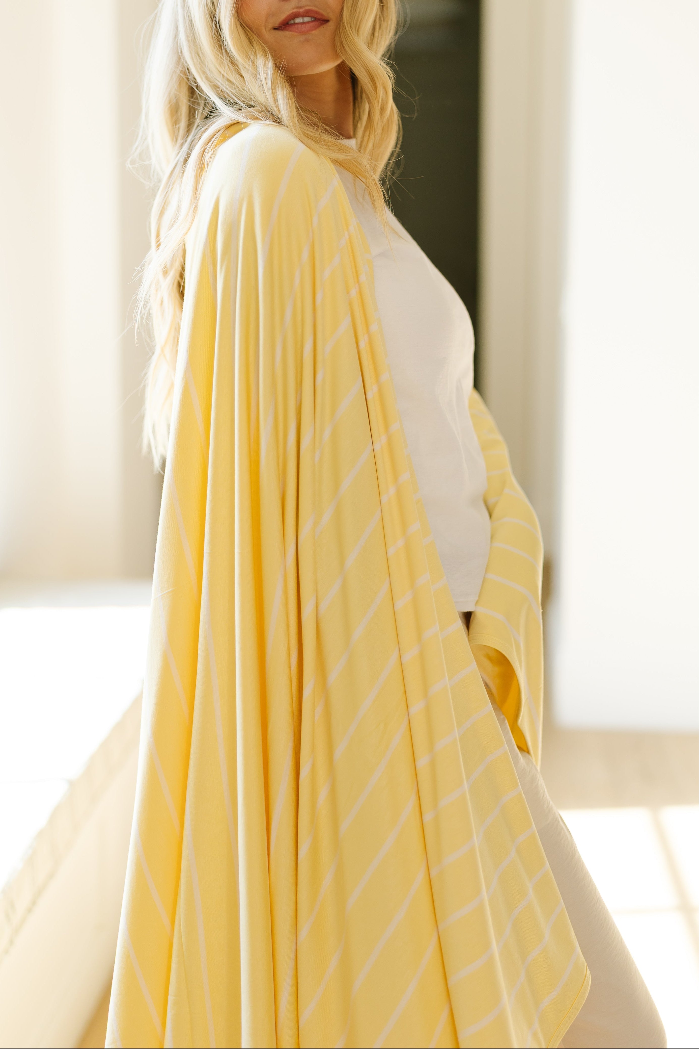 A woman with long blonde hair, dressed in a white top and wrapped in the yellow striped "lazy days" blanket, stands relaxed by a sunlit window, gazing over her shoulder.
