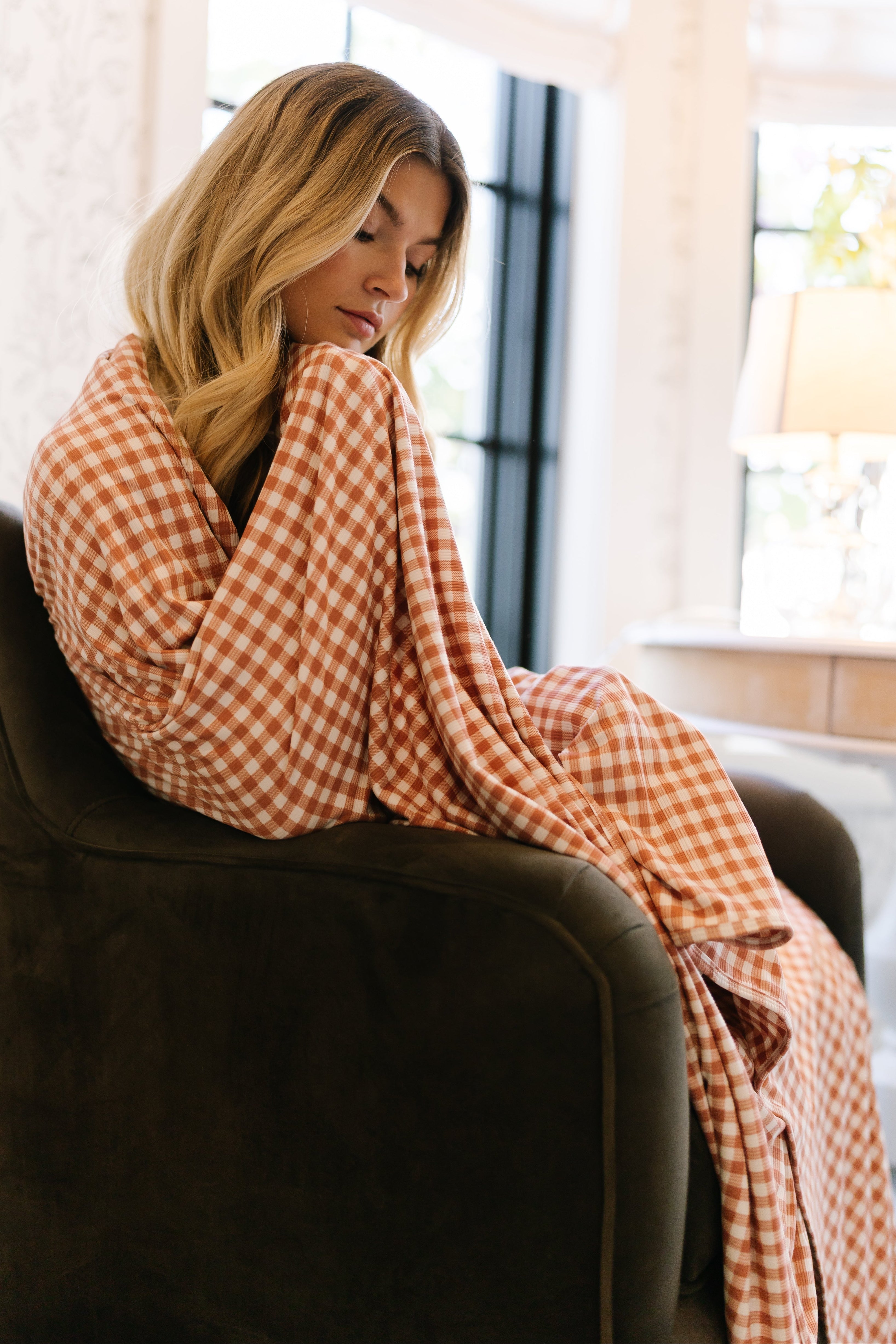 A woman with long blonde hair sits on a dark chair, wrapped in the dolly gingham check blanket. She gazes down thoughtfully in a bright room with large windows and a desk lamp.