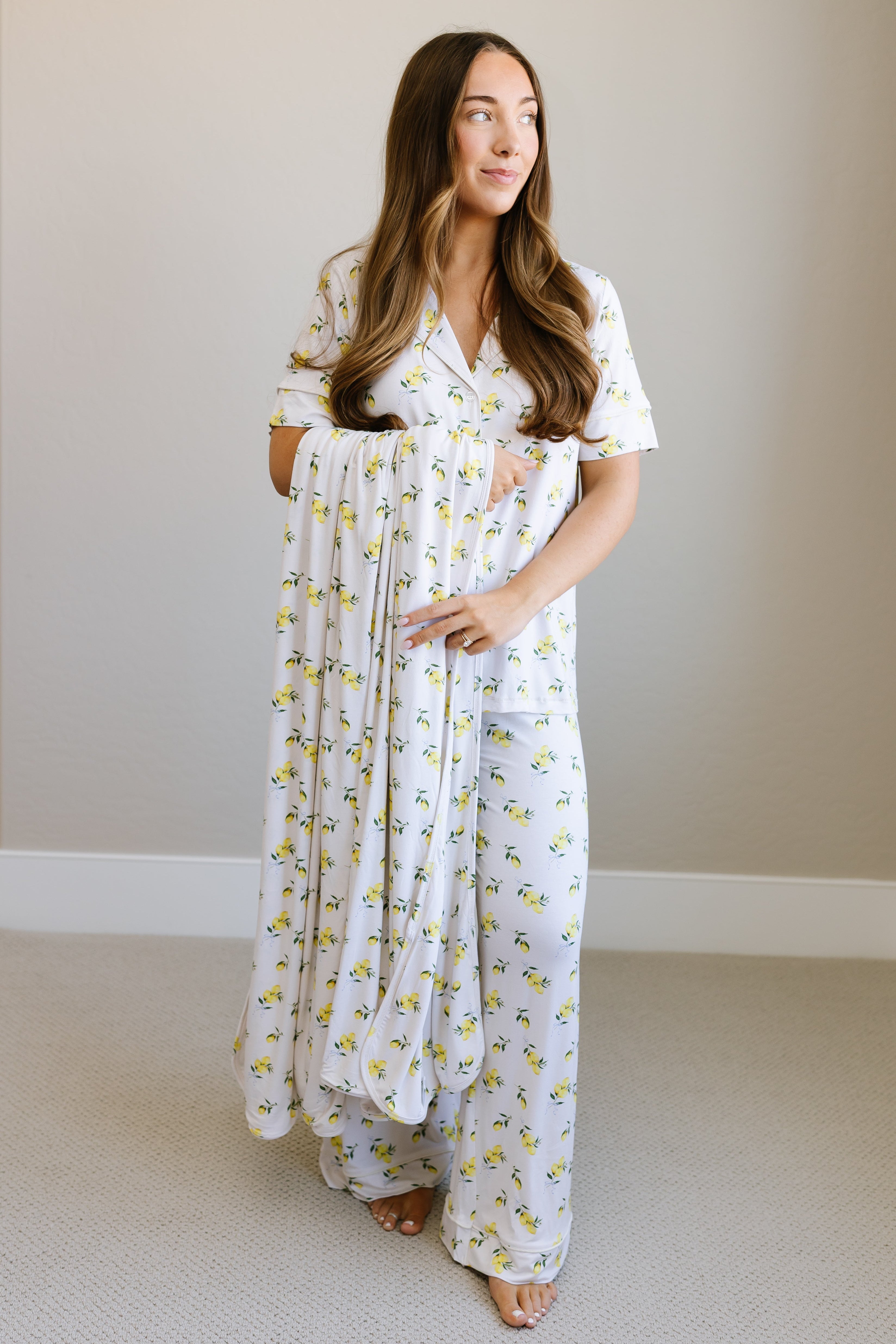 A woman with long brown hair stands indoors in matching limoncello pajamas with a yellow floral print, holding a baby wrapped in a luxurious limoncello bamboo blanket with the same pattern. Both face a window, softly lit by natural light.