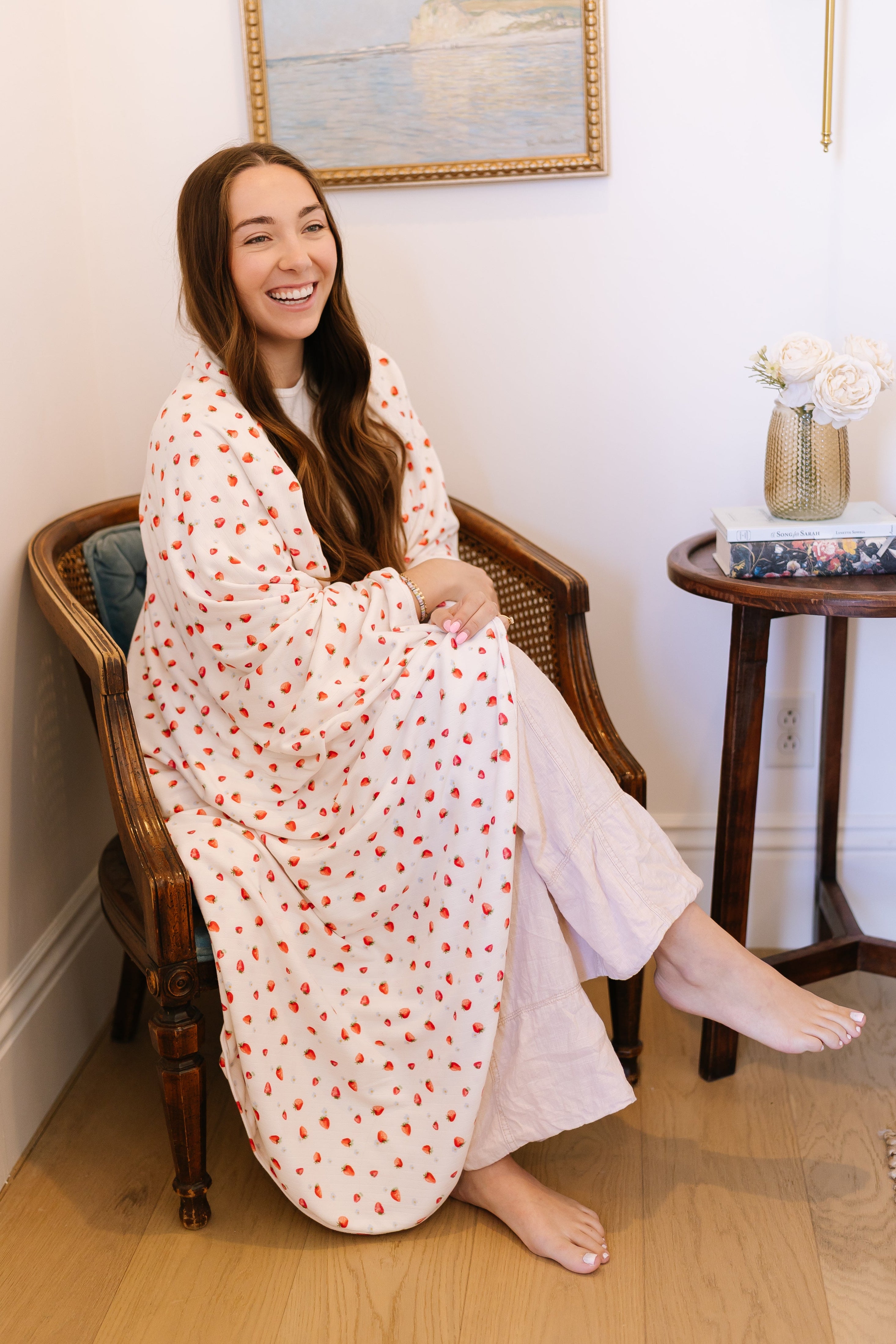 A woman with long brown hair smiles as she sits barefoot in a cozy, sunlit room, wrapped in the strawberry blossom / pointelle blanket—red polka dots add charm. By her side is a small table with flowers, a clock, and decor.