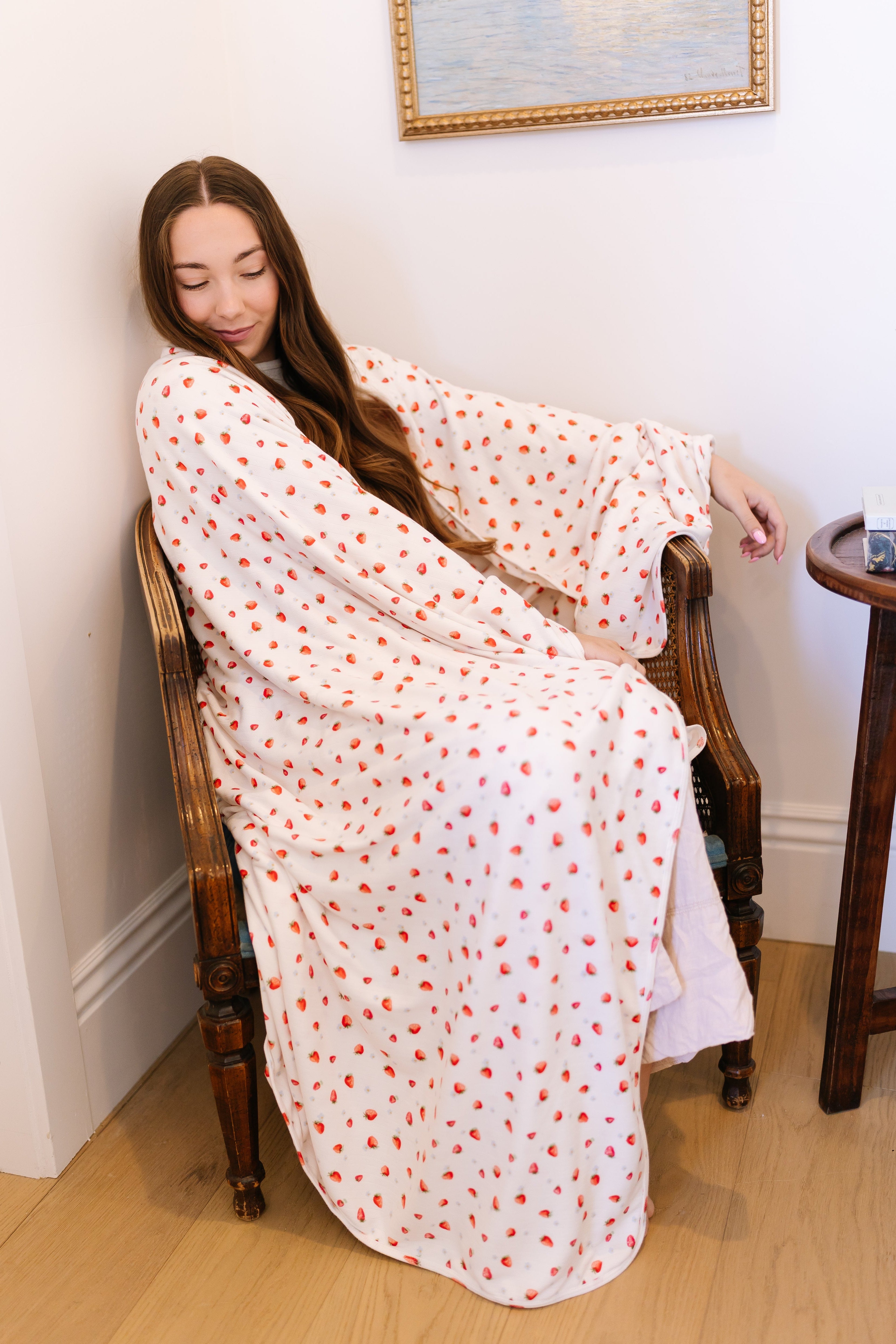 Relaxed with her eyes closed, a woman with long brown hair sits in a wooden chair, wearing the strawberry blossom / pointelle robe. Beside her are a small round table and a framed seascape painting.