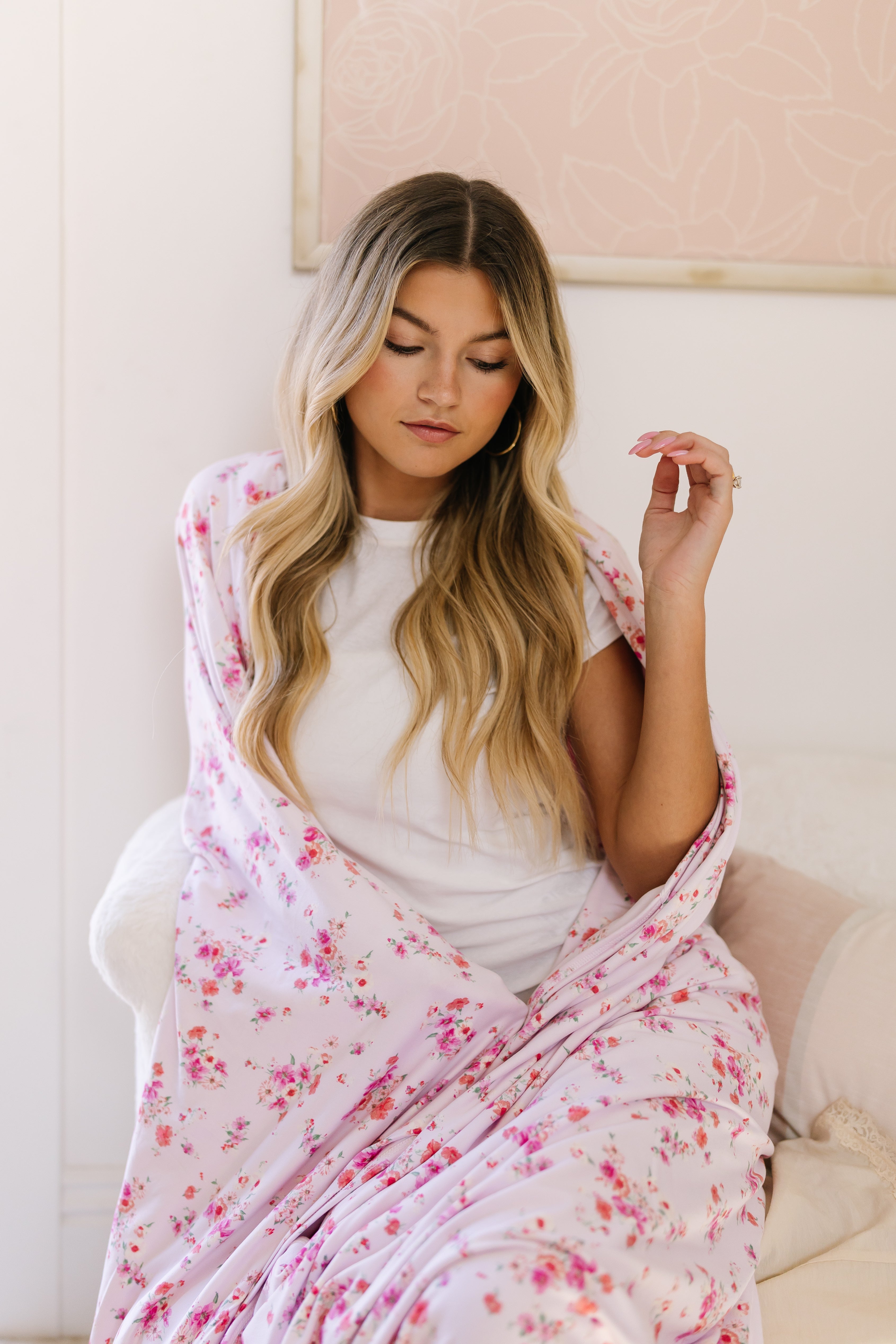 A woman with long, wavy blonde hair sits indoors, wrapped in the extra soft pink floral "sweet bloom" blanket. She wears a white t-shirt and looks down with a relaxed expression, her hand gently raised near her face.
