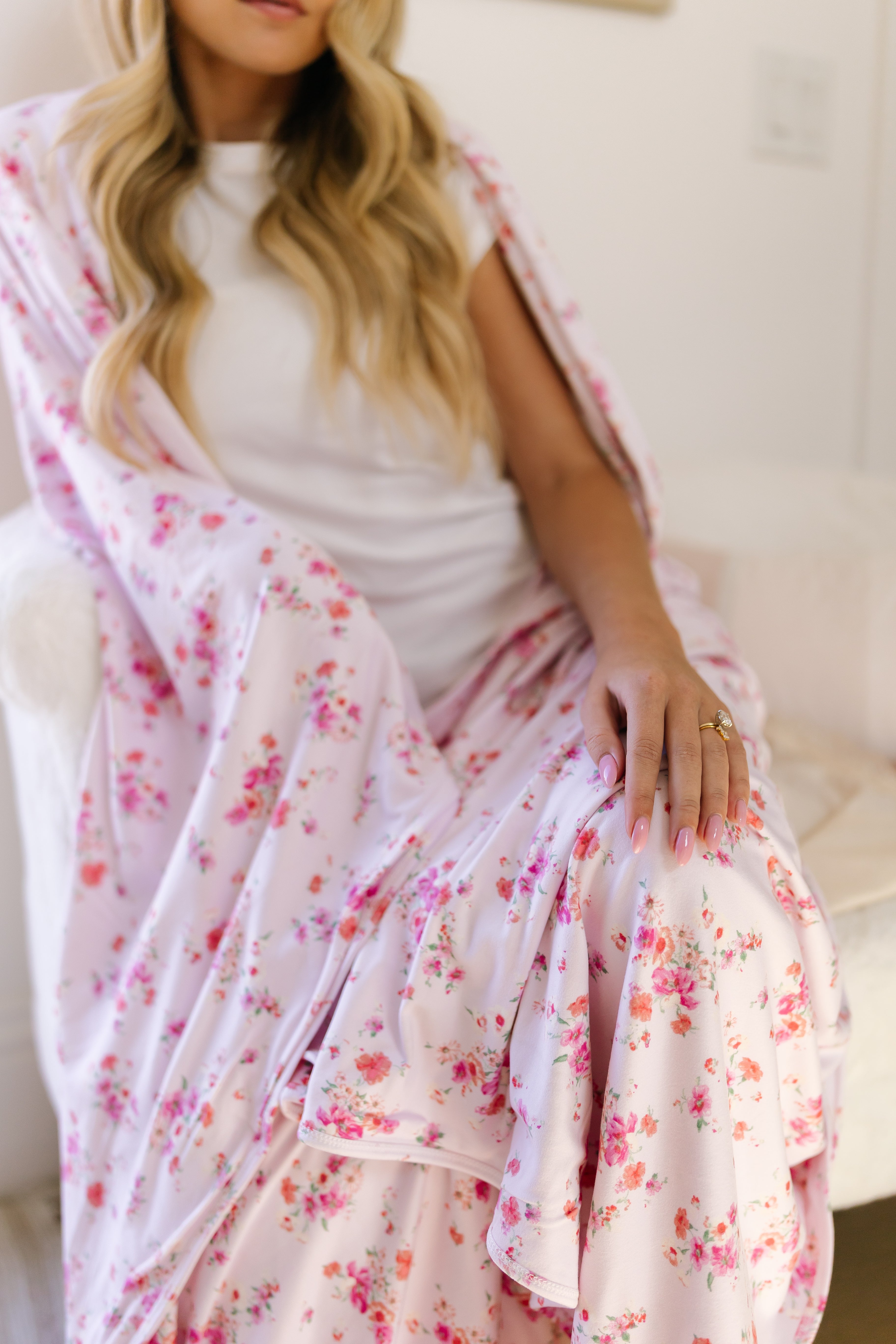 A woman with long blonde hair sits in a chair, wearing a white top with the "sweet bloom" pink floral blanket draped over her lap. Her hand rests on the extra soft "sweet bloom" blanket, showing off a ring on her finger.