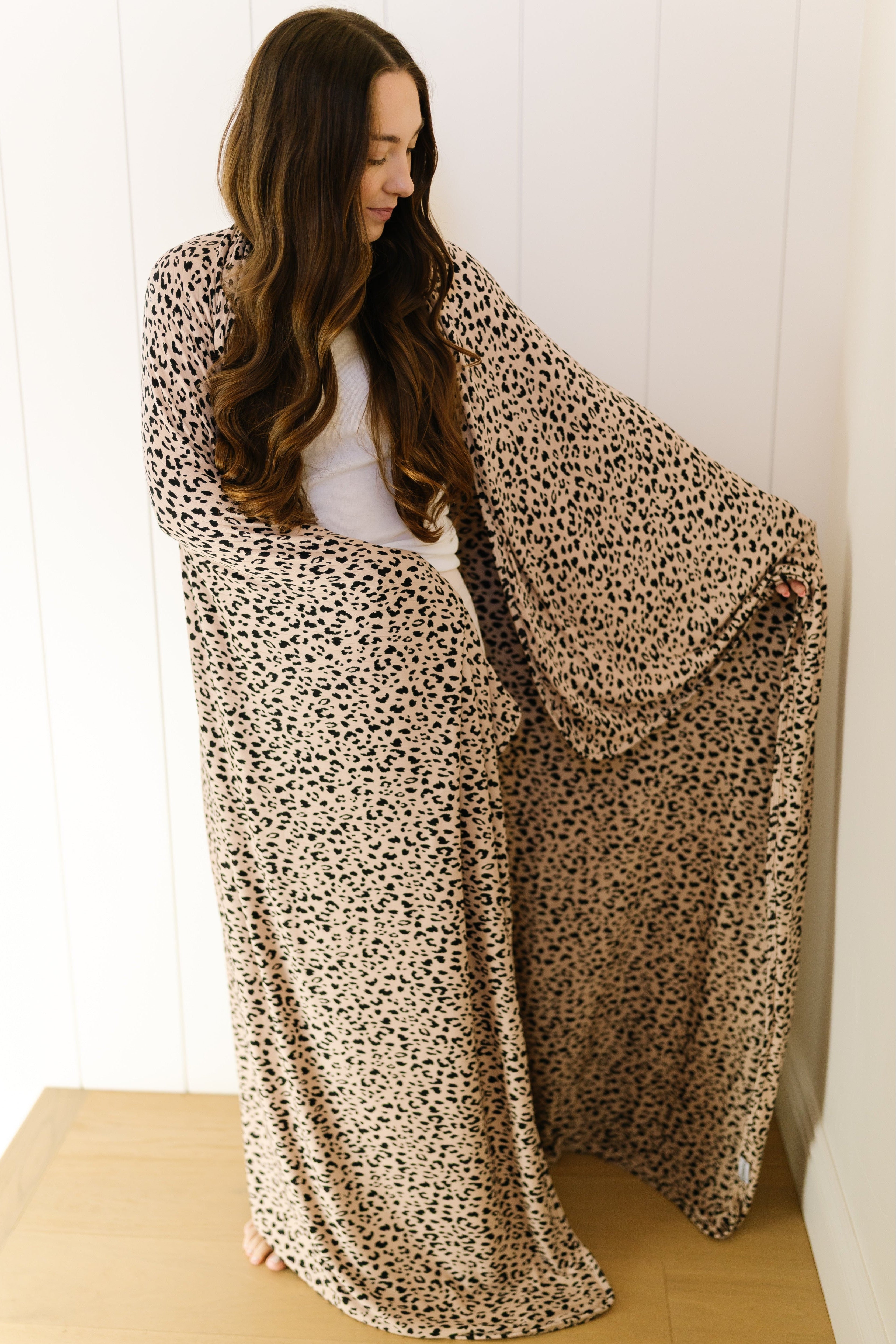 A woman with long, wavy brown hair wears the "cheetah girl / pointelle" robe with wide sleeves, standing barefoot on a light wooden floor against a white paneled wall, looking down and holding out one side like a luxurious blanket.