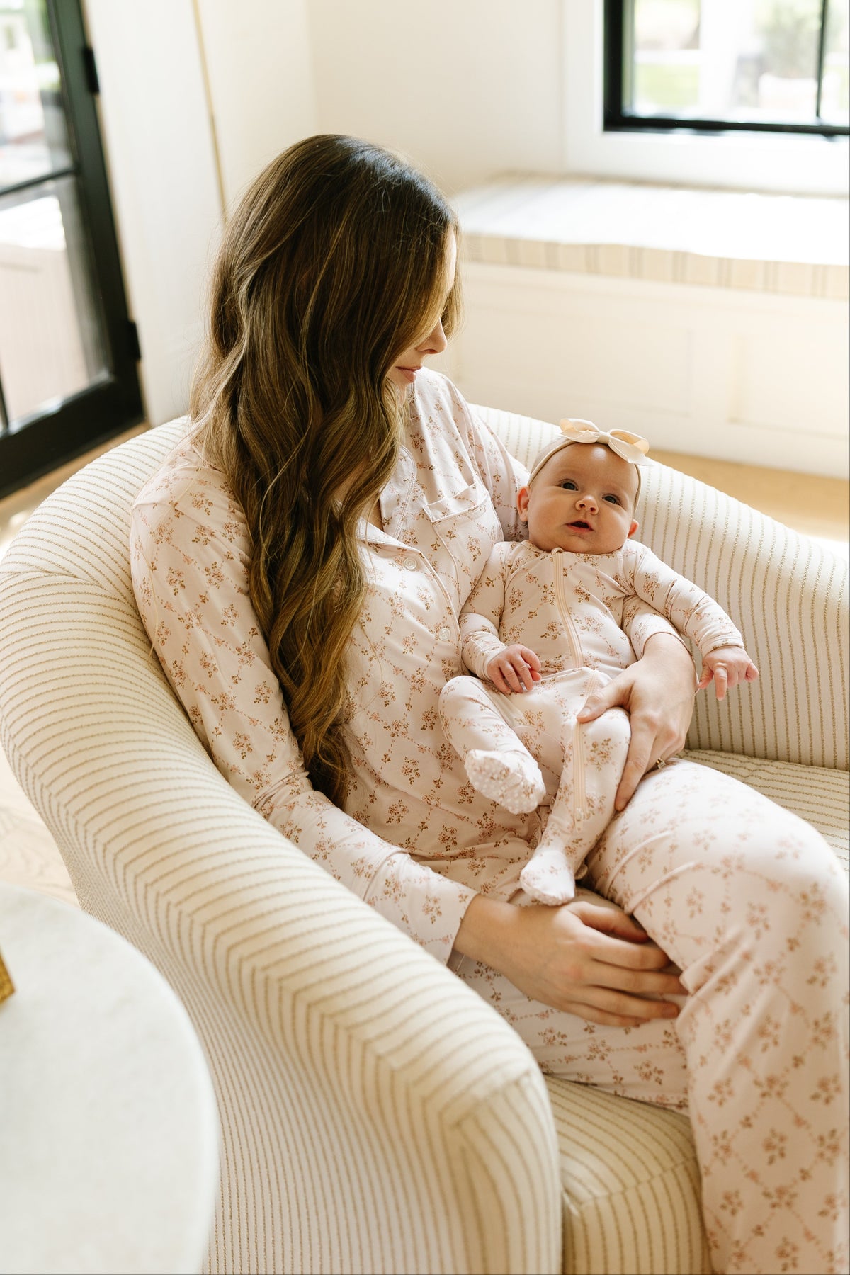 A woman with long hair sits in a striped armchair, holding a baby dressed in a cozy keepsake pajama set with long pants and matching floral print sleepwear, complete with an adorable headband. They are indoors near a window, sharing a quiet, tender moment.