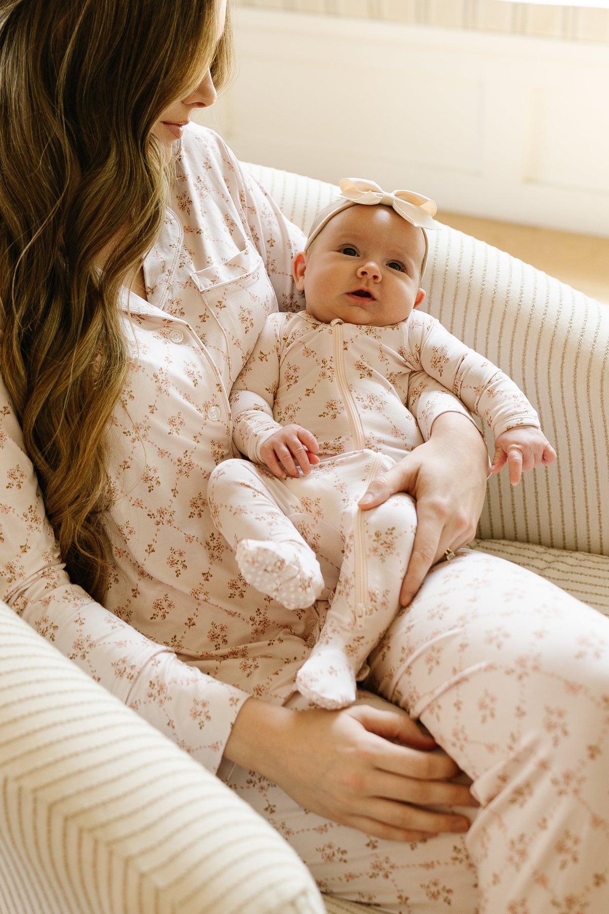 A woman sits with a baby in her lap on a striped chair. Both are wearing matching light-colored pajamas with delicate floral prints. The baby is dressed in the cozy keepsake one piece zipper and wears a bow headband, gazing upward as the woman’s face is partially seen.