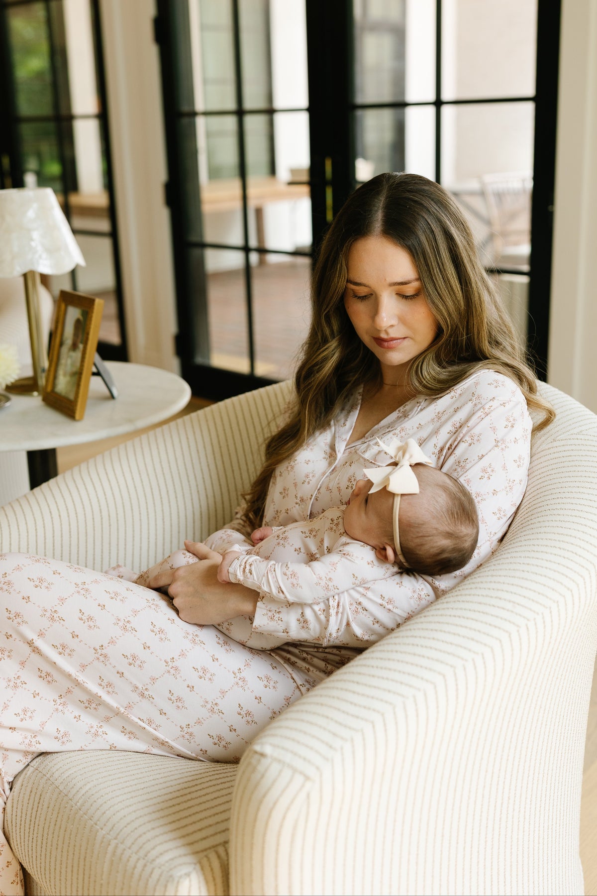 A woman wearing the keepsake pajama set with long pants holds a baby with a bow headband while sitting in a striped armchair near a table and large glass doors.