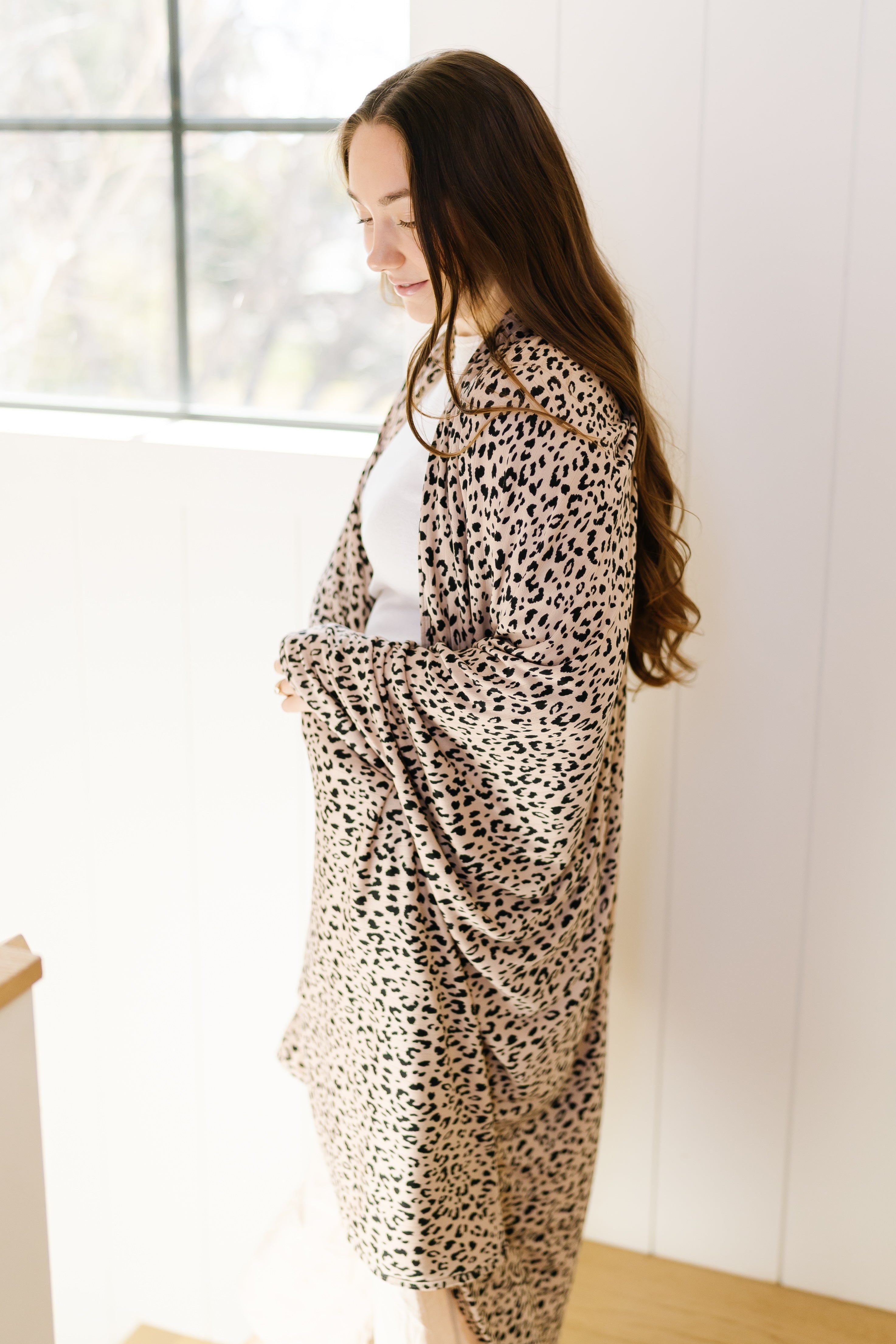 A woman with long brown hair stands indoors by a window, wearing a white top and the cheetah girl / pointelle blanket draped over her shoulders. Soft natural light filters in, creating a calm and cozy atmosphere.