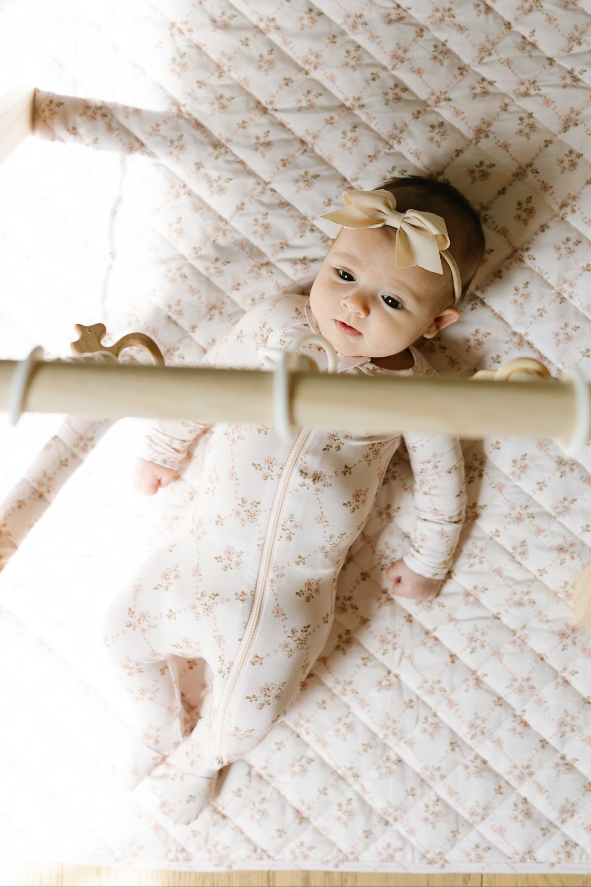 A baby wearing a keepsake / one piece zipper lies on a patterned quilt, looking up at a wooden play gym with hanging toys.