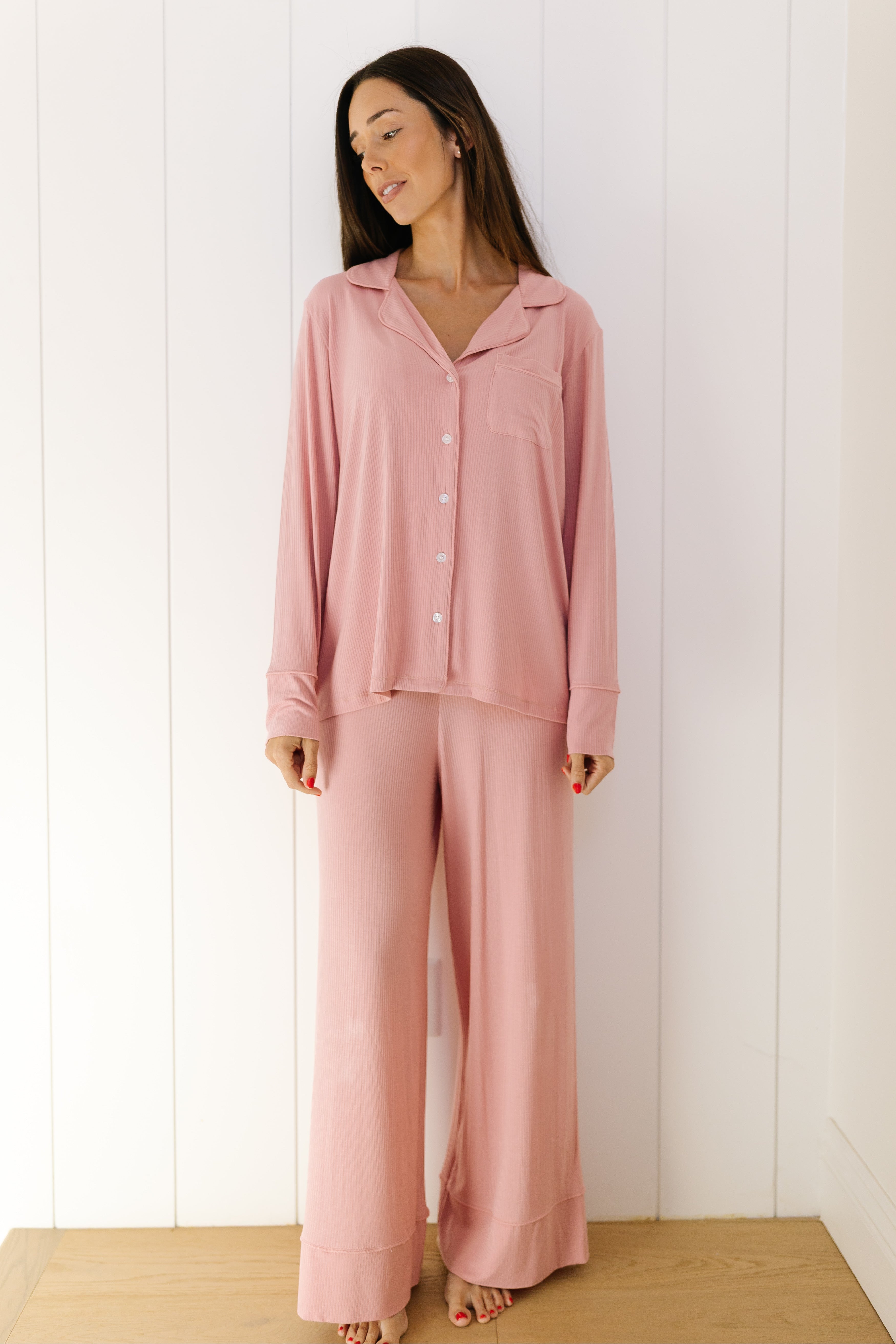 A woman with long dark hair stands indoors against a white wall, smiling slightly as she wears the peony pajama set—a light pink women's lounge set featuring a button-up top and wide-leg long pants.
