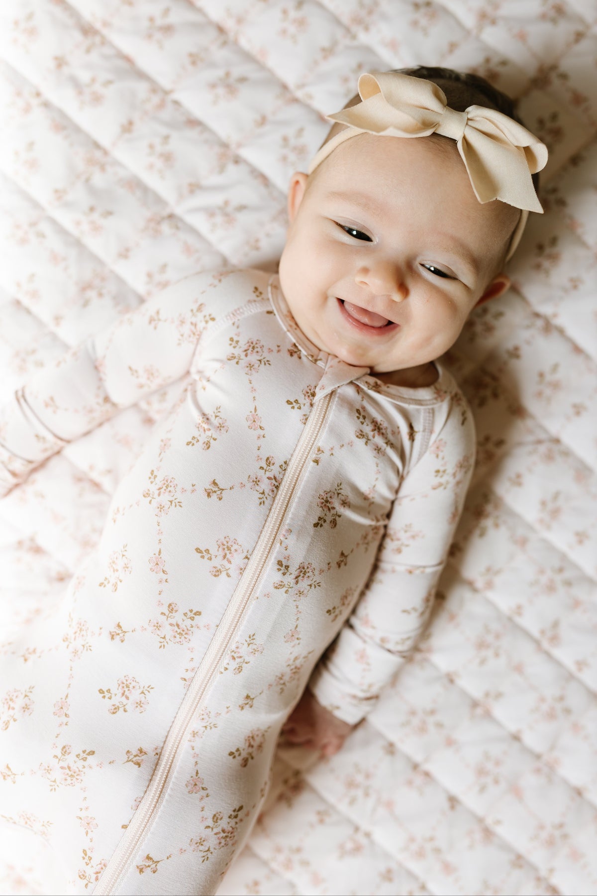 A smiling baby wears the keepsake / one piece zipper, complete with a big bow headband, lying on a quilted blanket with a matching floral pattern—perfectly cozy in this soft bamboo sleepwear for ultimate comfort.
