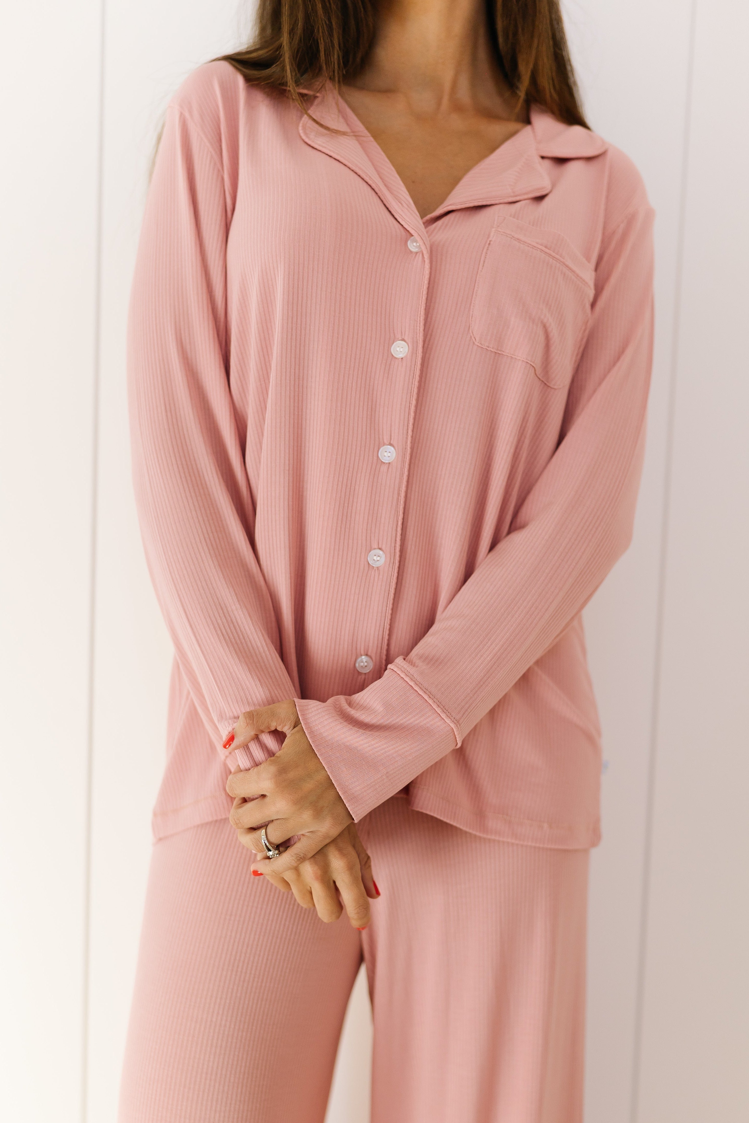 A woman stands against a white wall, arms gently crossed, wearing the peony pajama set with a light pink long-sleeve button-up top and matching loose long pants. Her head is cropped out of the frame.