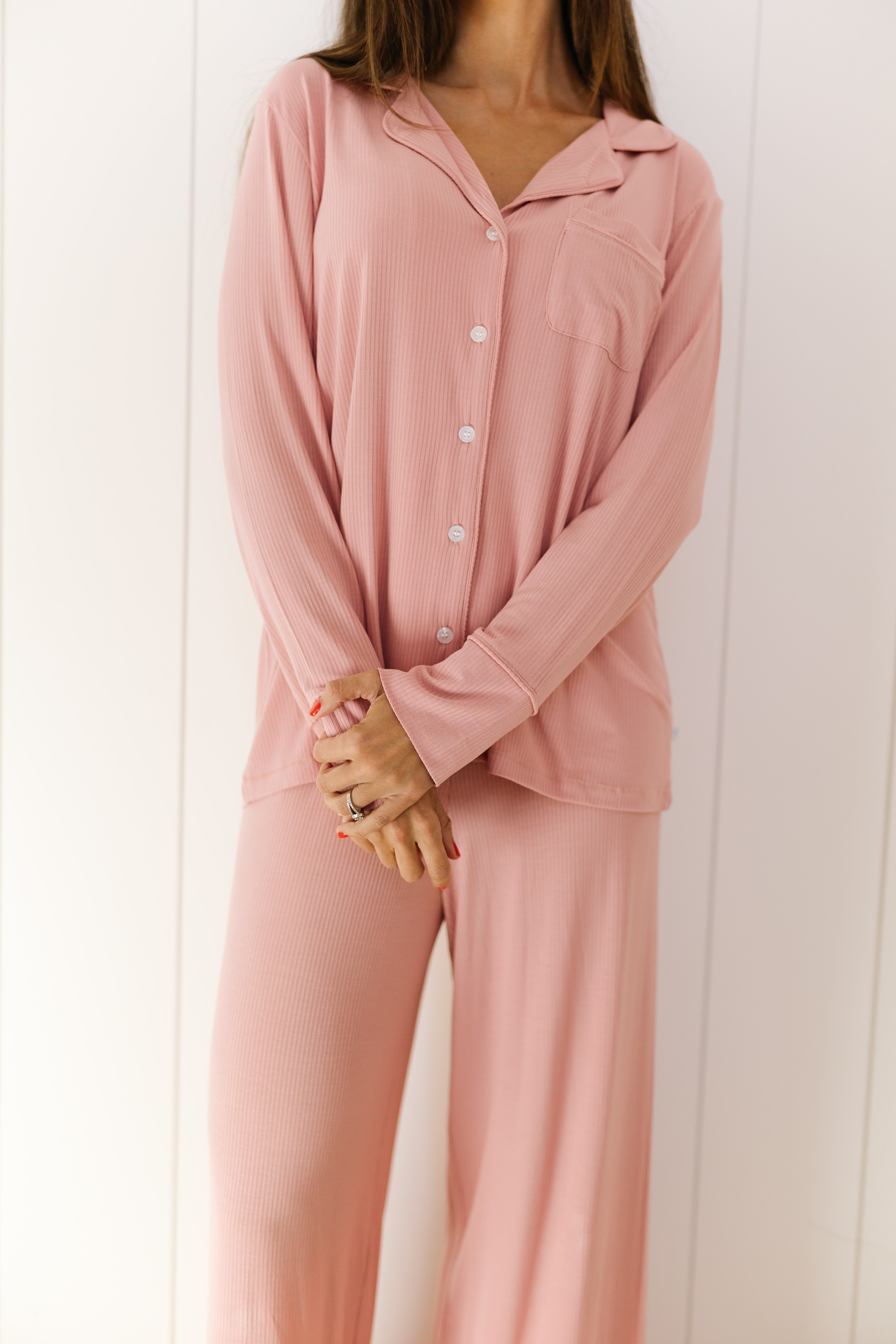A woman stands against a white wall, arms gently crossed, wearing the peony pajama set with a light pink long-sleeve button-up top and matching loose long pants. Her head is cropped out of the frame.