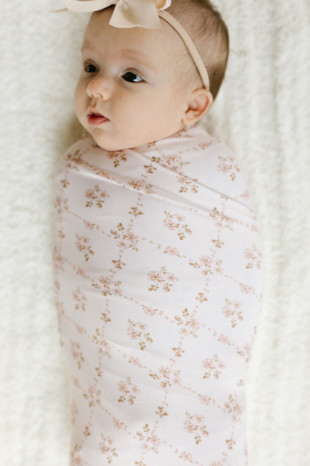 A baby in the keepsake / swaddle, featuring a light pink floral print, is lying on a soft white surface and wearing a headband with a bow while gazing off to the side.