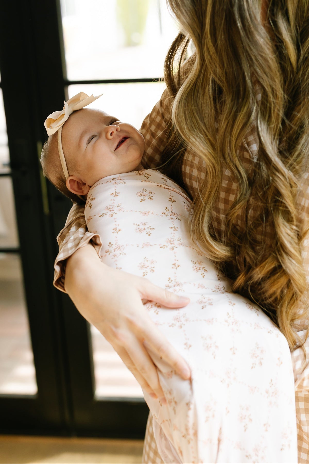 A woman with long wavy hair, wearing a gingham shirt, holds a baby wrapped in the keepsake / swaddle featuring a cozy floral print and a bow headband, standing in front of glass doors.