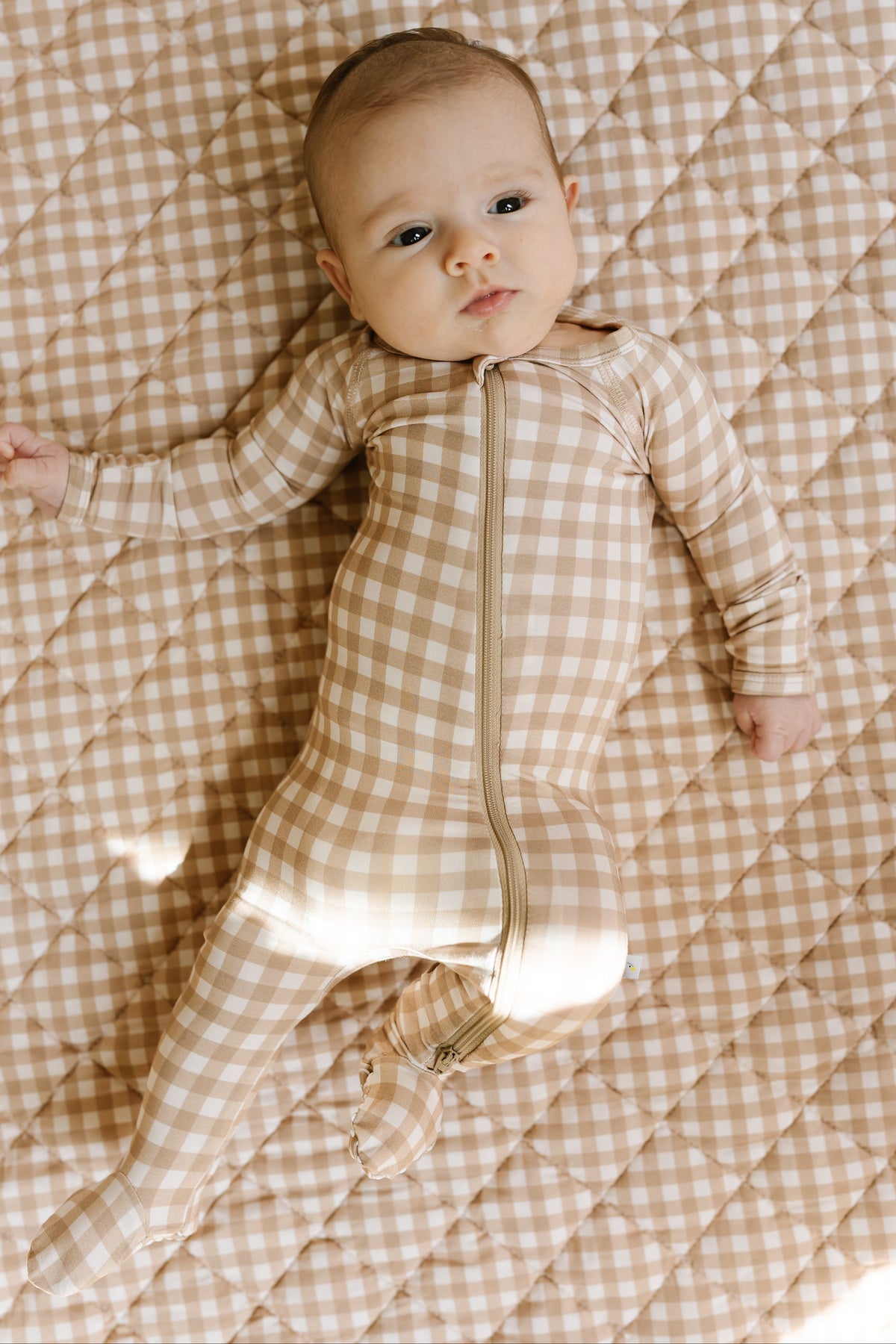 A baby lies on a beige and white checkered “teddy bear check / one piece zipper” outfit, looking up with a calm expression.