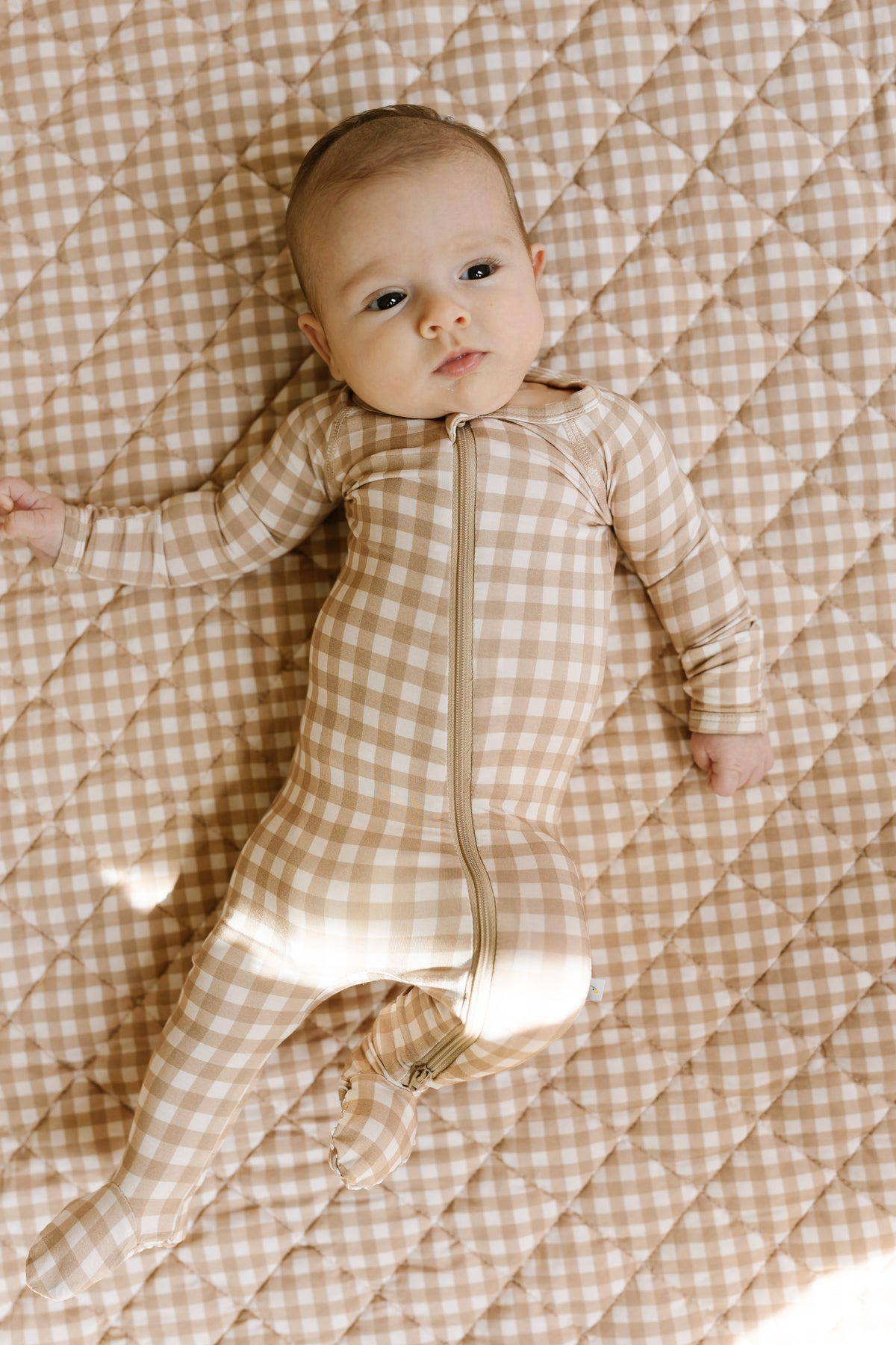 A baby lies on a beige and white checkered “teddy bear check / one piece zipper” outfit, looking up with a calm expression.