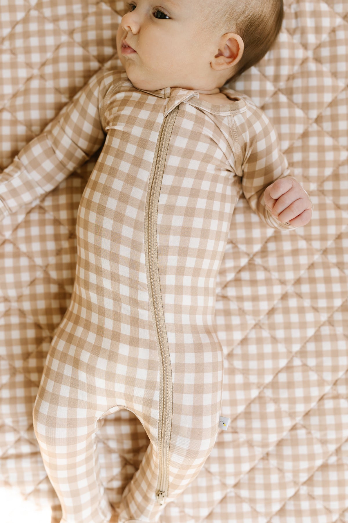 A baby lies on a quilted blanket, wearing the teddy bear check / one piece zipper, and looks to the side.