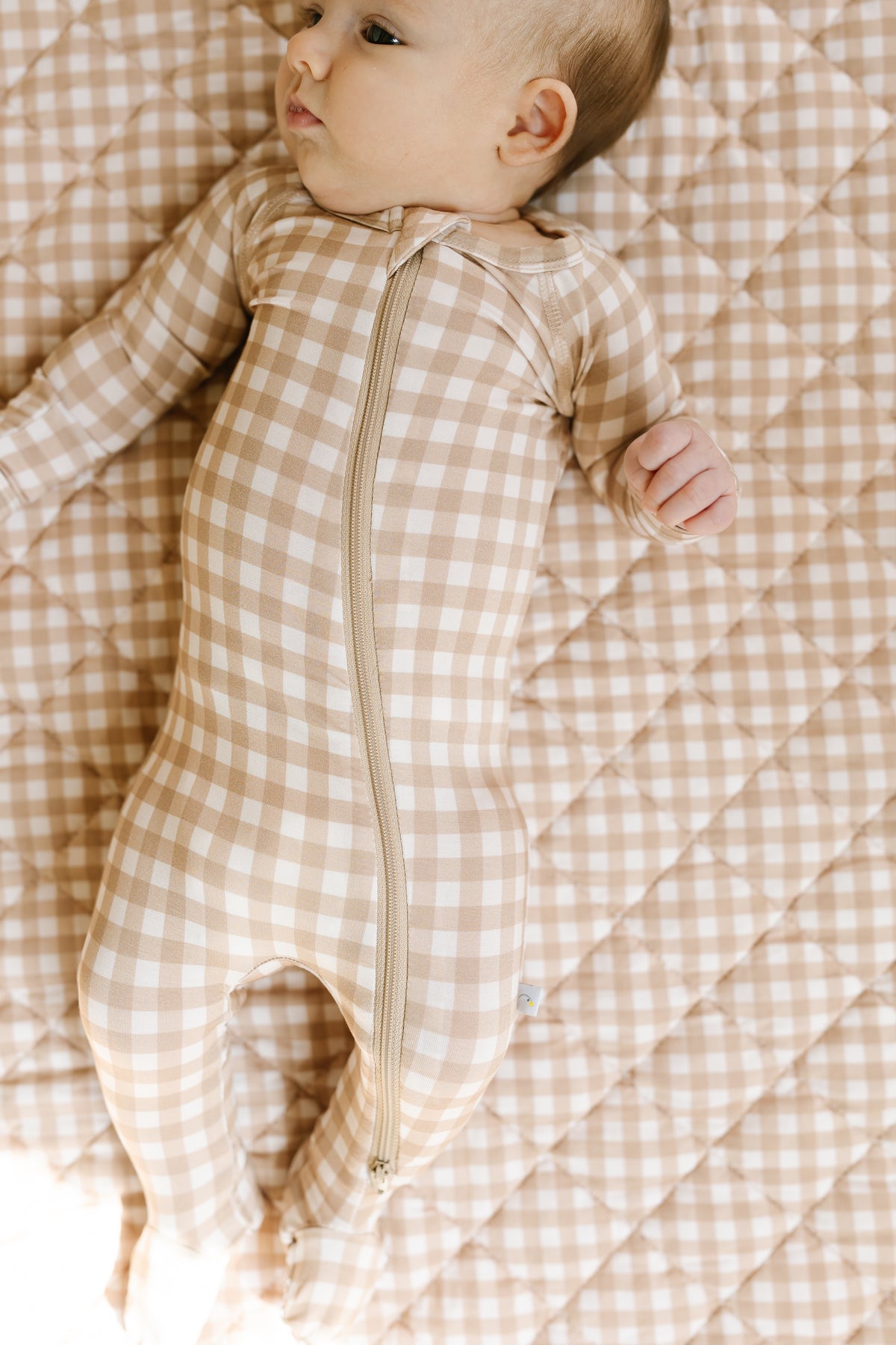 A baby lies on a quilted blanket, wearing the teddy bear check / one piece zipper, and looks to the side.