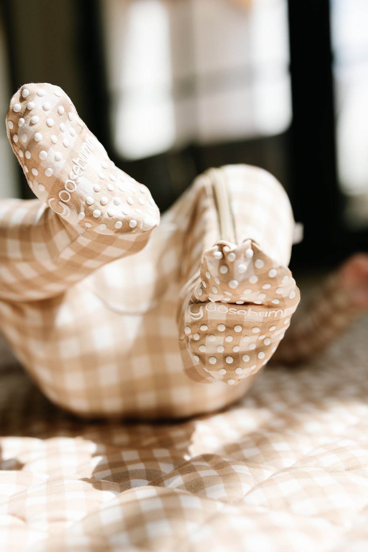 A baby in the teddy bear check / one piece zipper, featuring beige and white checkered fabric and textured polka-dot soles, lies on a matching surface with legs playfully kicking up.