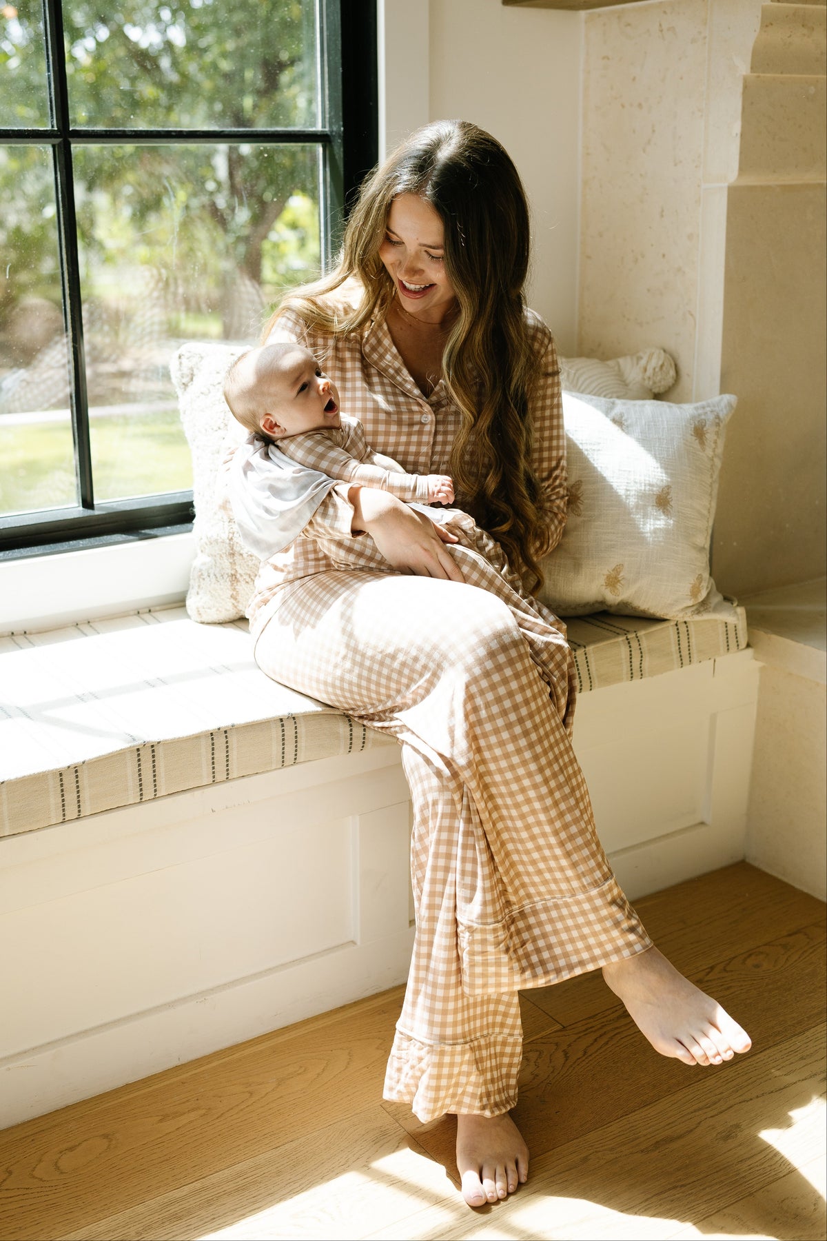 A woman in a teddy bear check pajama set with long pants sits barefoot on a window seat, smiling and holding a baby who gazes up at her. Sunlight streams through the window, illuminating the cozy scene and highlighting the relaxed fit loungewear that adds to the tranquil atmosphere.