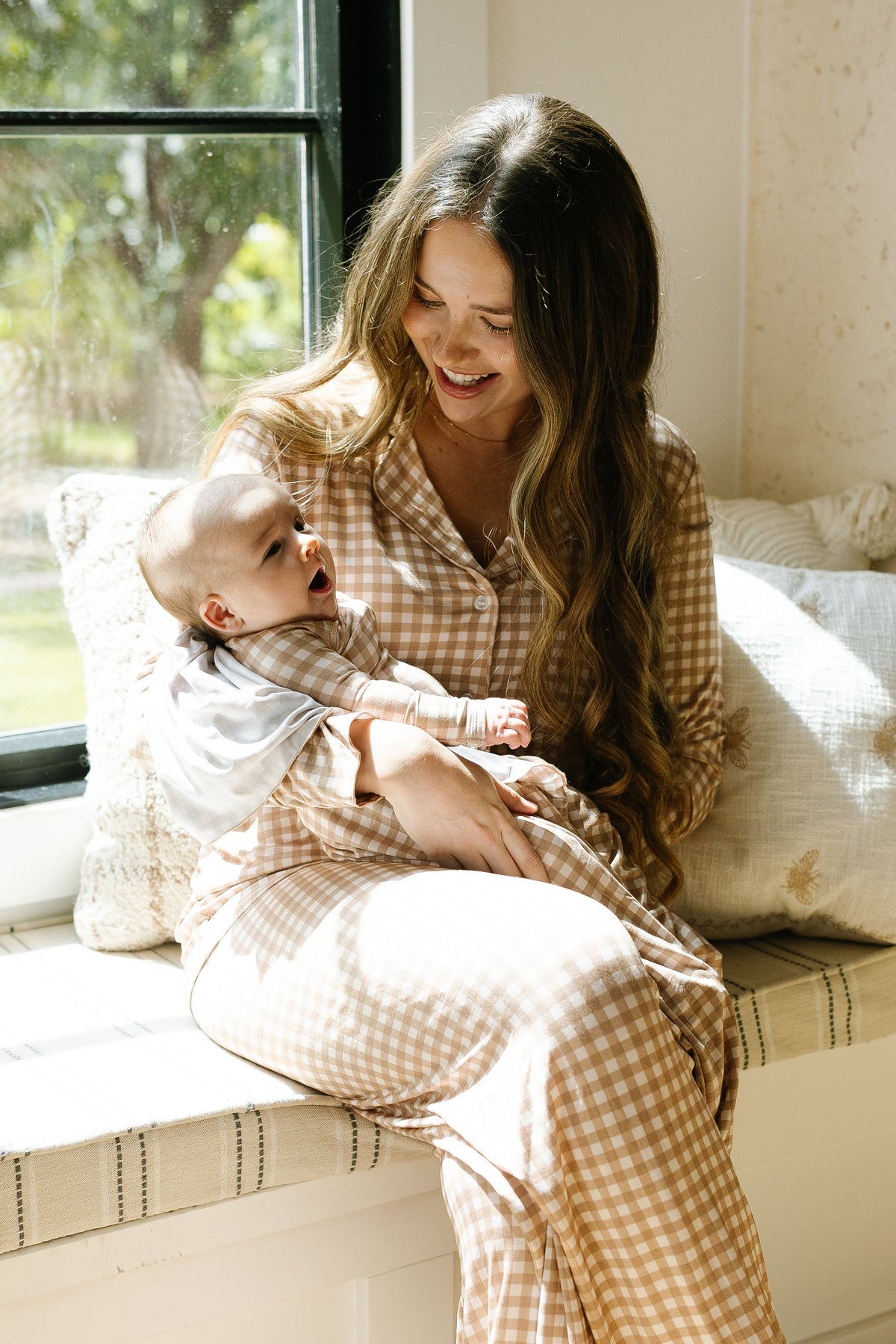 A woman in teddy bear check pajamas sits by a window, smiling at a baby in her lap dressed in a soft teddy bear check one piece zipper. Sunlight streams in, creating a warm, cozy atmosphere.