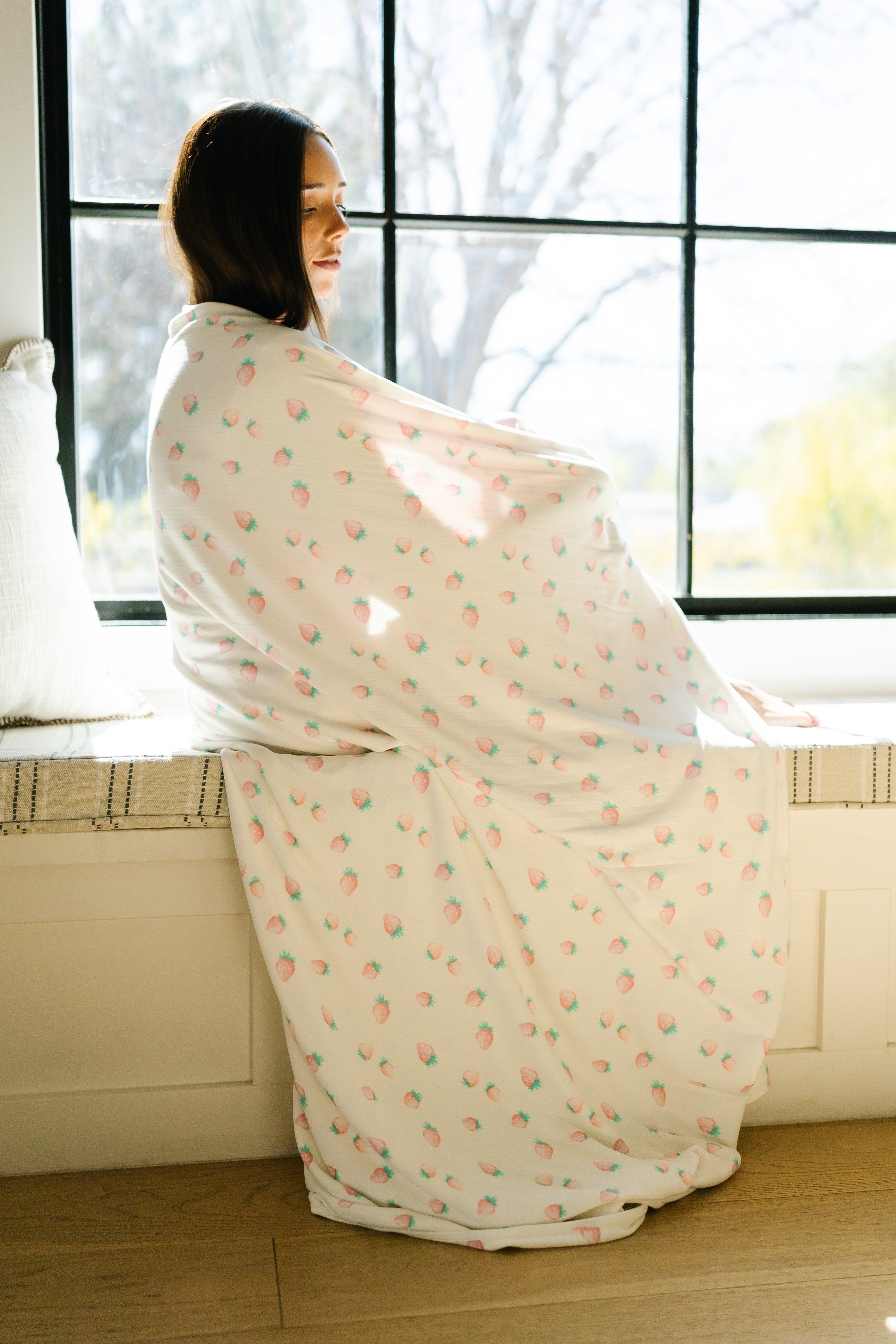 A woman sits on a window seat wrapped in the pink berry / pointelle blanket, smiling softly as sunlight streams through the large window behind her.