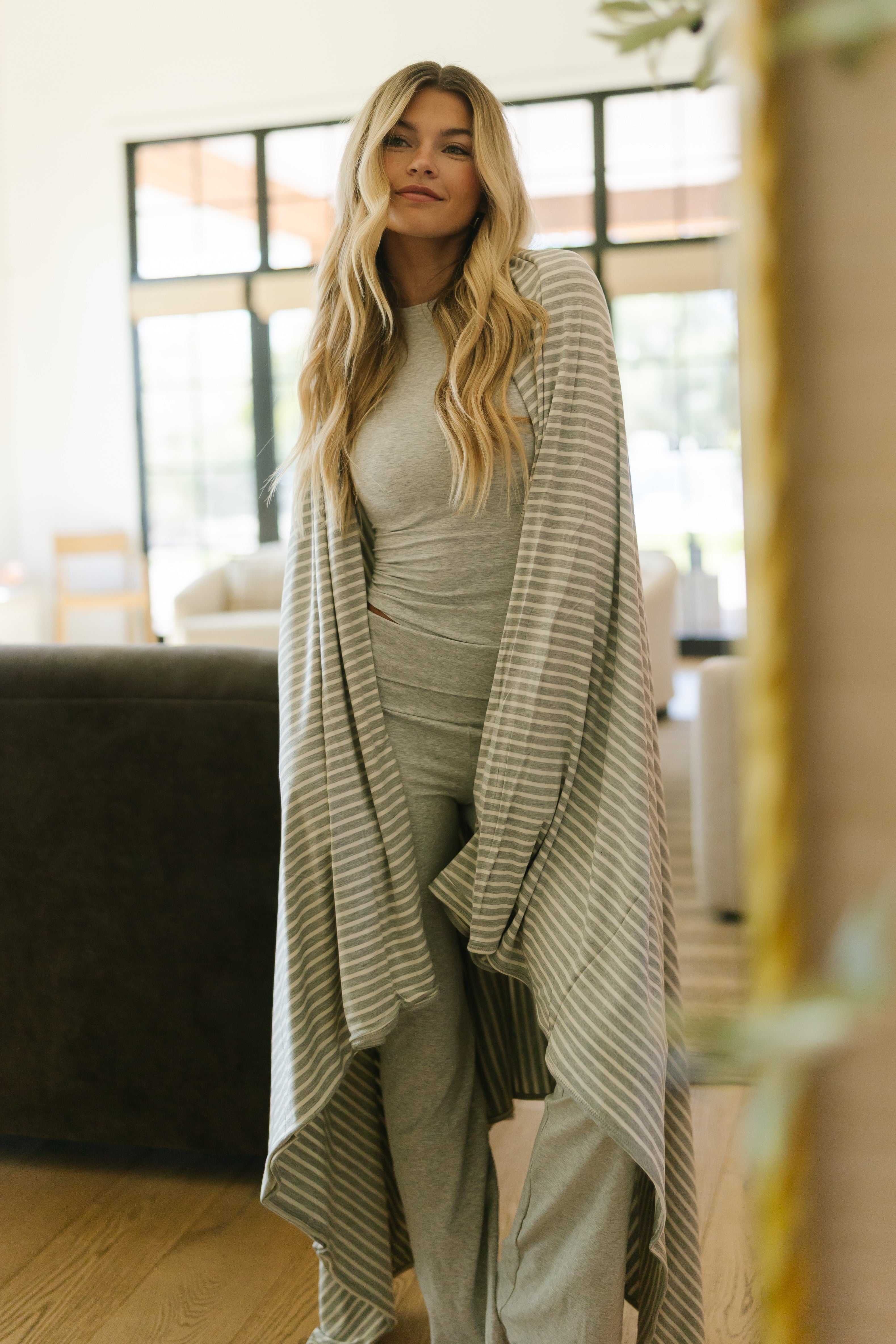 A woman with long blonde hair stands indoors wearing the basically / heather loungewear set and a grey striped cardigan, wrapped in a modern blanket, looking relaxed in a bright living room with large windows.