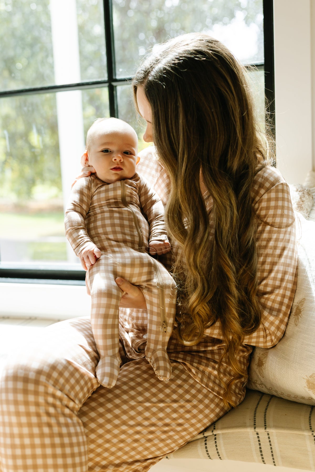 A mother with long brown hair, wearing matching tan and white pajamas, sits by a window and holds her young baby, who is dressed in a teddy bear check one piece zipper.