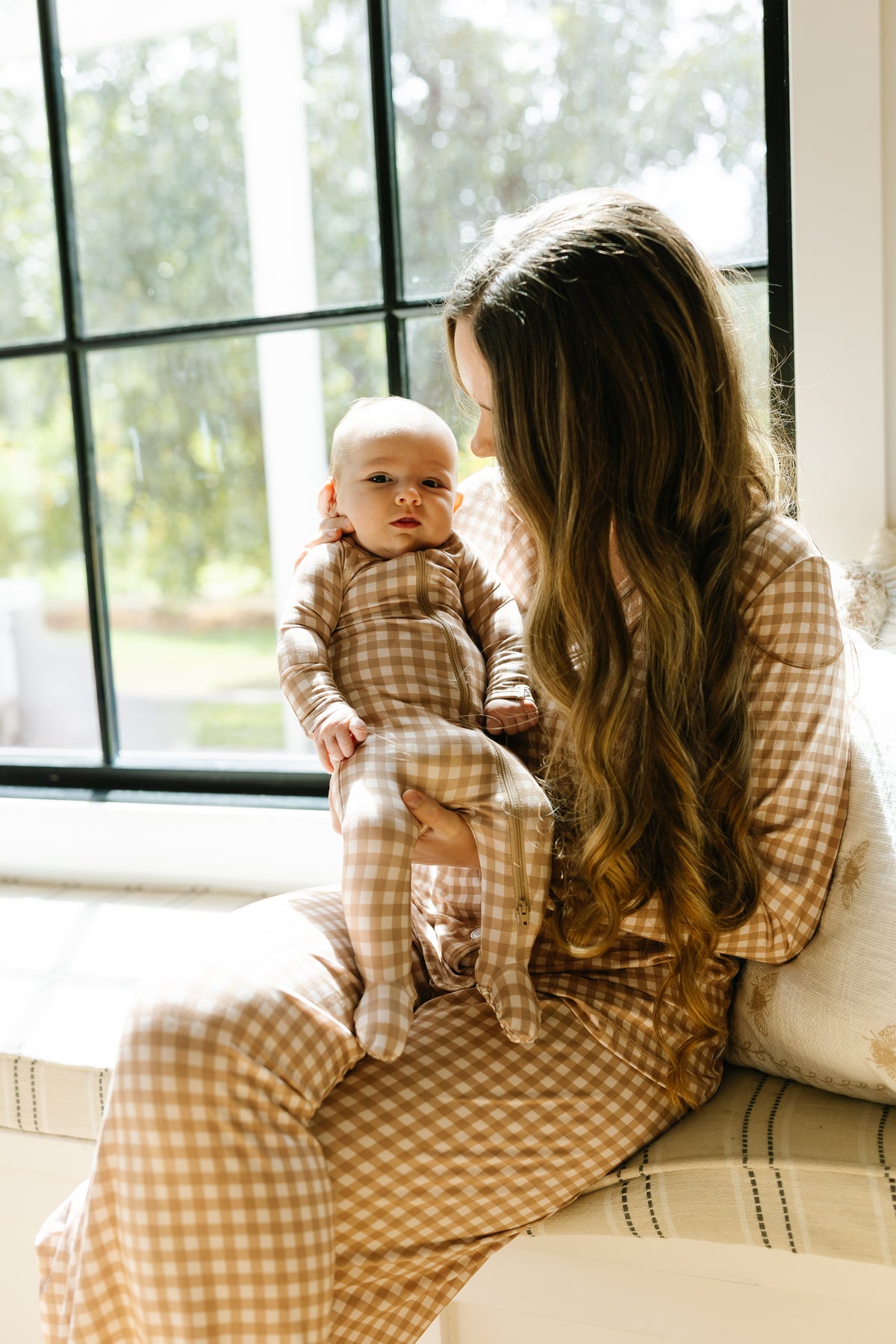 A mother with long brown hair, wearing matching tan and white pajamas, sits by a window and holds her young baby, who is dressed in a teddy bear check one piece zipper.