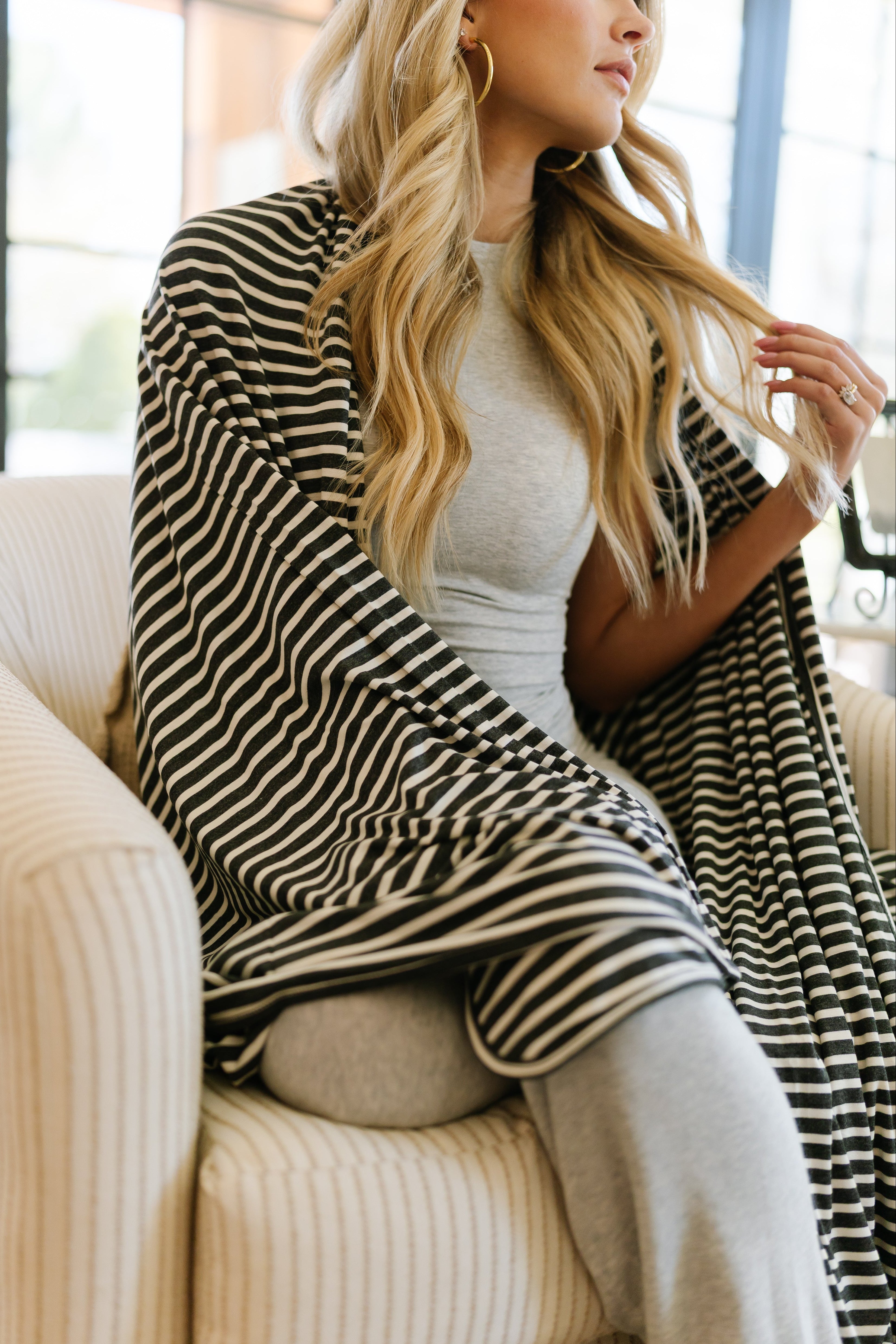 A woman with long blonde hair sits in a beige armchair, wrapped in the basically / charcoal modal rayon blanket over her light gray outfit and black-and-white striped shawl, gently holding a strand of her hair.