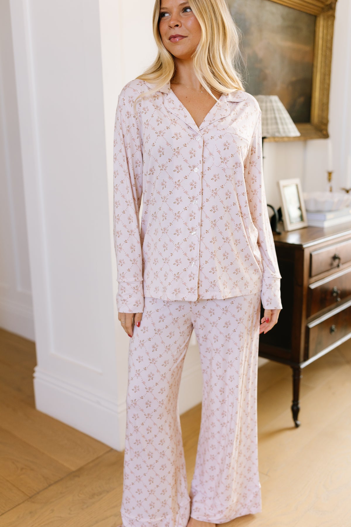 A woman stands indoors wearing the keepsake long pant pajama set, featuring a light pink floral print. She has blonde hair and is smiling slightly. Behind her is a wooden dresser with a lamp, photo frame, and art on the wall.