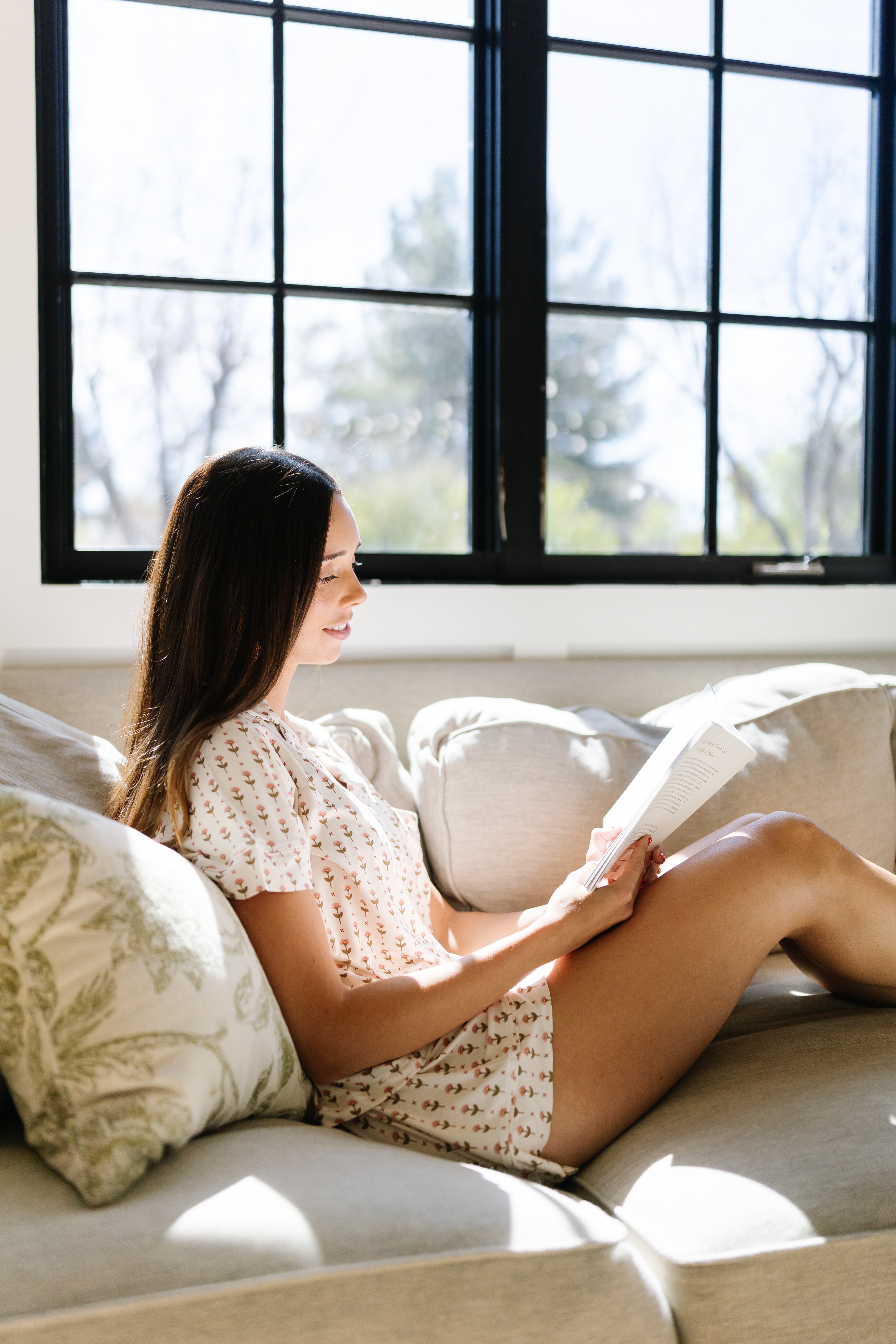 A woman wearing the tulip pajama set with shorts sits on a light-colored sofa by a window, reading a book in natural sunlight. The bright, cozy room features decorative pillows.