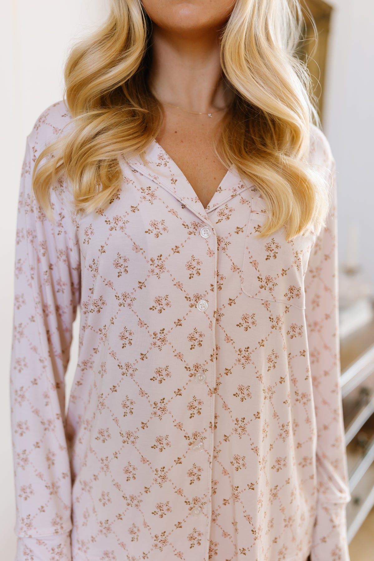 A woman with wavy blonde hair wears the keepsake pajama set, featuring a light pink long-sleeve button-up top adorned with a delicate floral pattern, embodying the comfort and elegance of women’s loungewear.