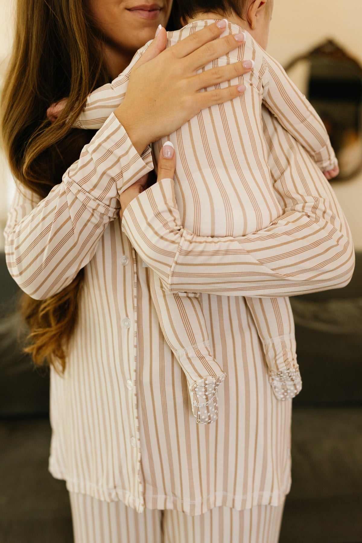 A woman with long hair holds a baby over her shoulder. Both are dressed in matching toast and honey pajama sets with long pants, featuring beige and white stripes and a relaxed fit. The baby’s face is not visible.