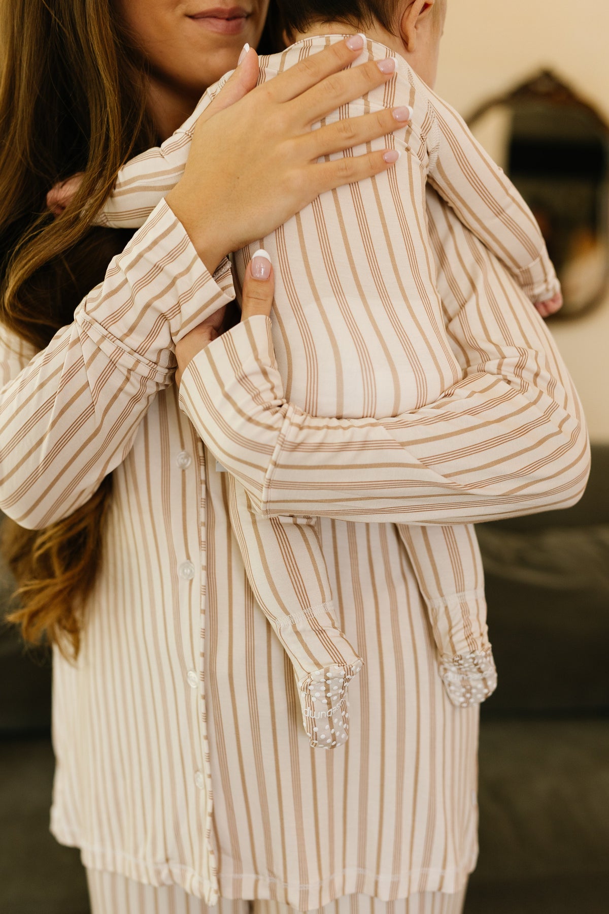 A woman holds a baby on her shoulder, both dressed in matching toast and honey / one piece zipper pajamas featuring beige and white stripes. Only the lower part of the woman’s face and the back of the baby’s head are visible, drawing attention to the soft comfort of their bamboo newborn pajamas.