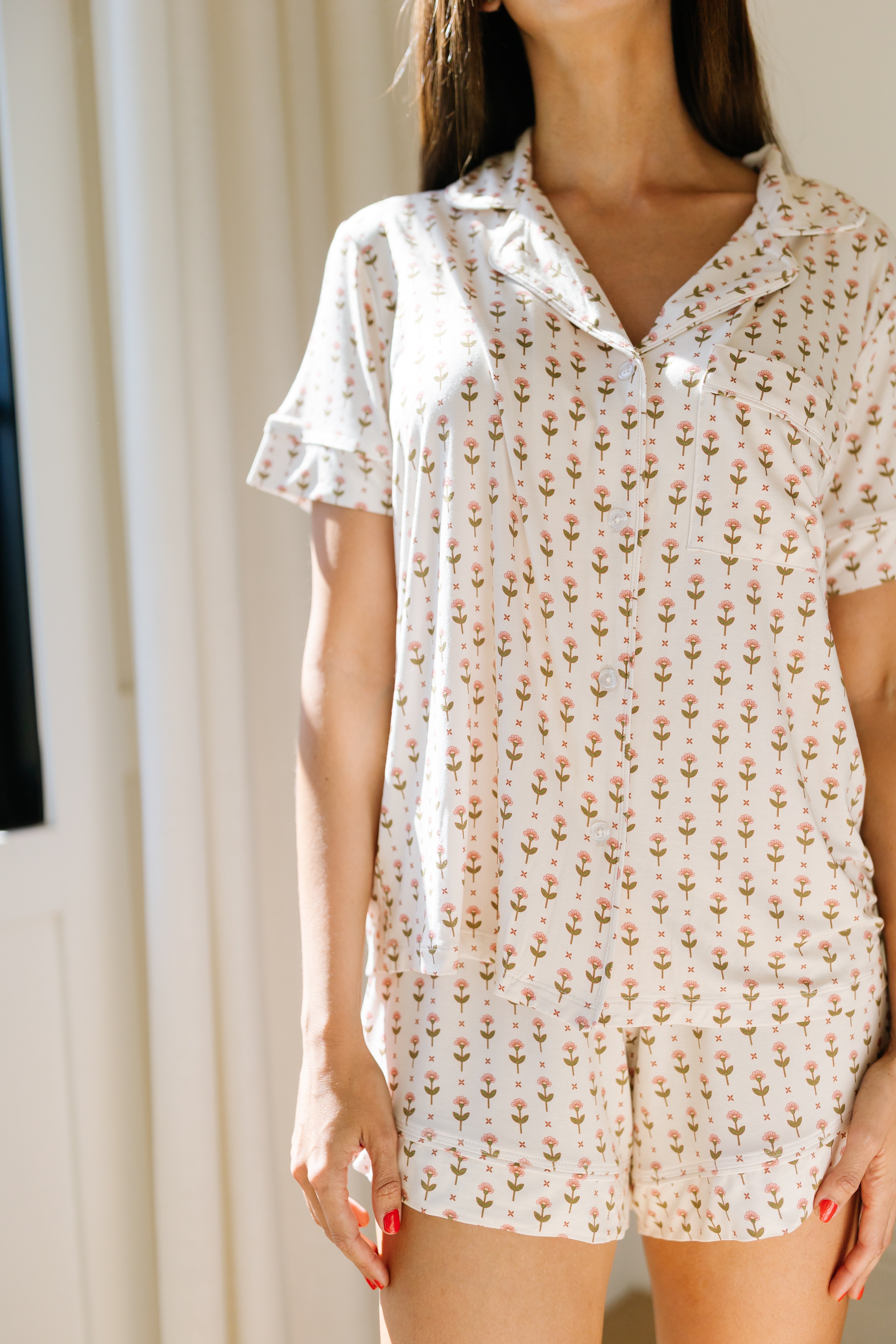 A woman indoors wears the tulip pajama set with shorts, featuring light-colored fabric, short sleeves, a small floral pattern, and a relaxed spring loungewear fit as sunlight streams in. Her face is partly out of frame.